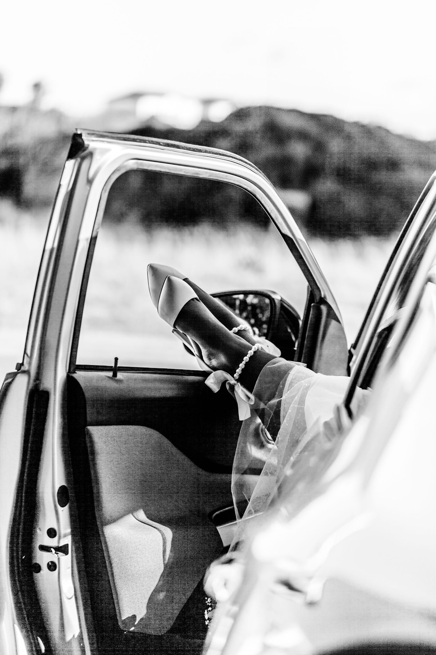 Black and white photo of bride in car with wedding dress visible through open door.