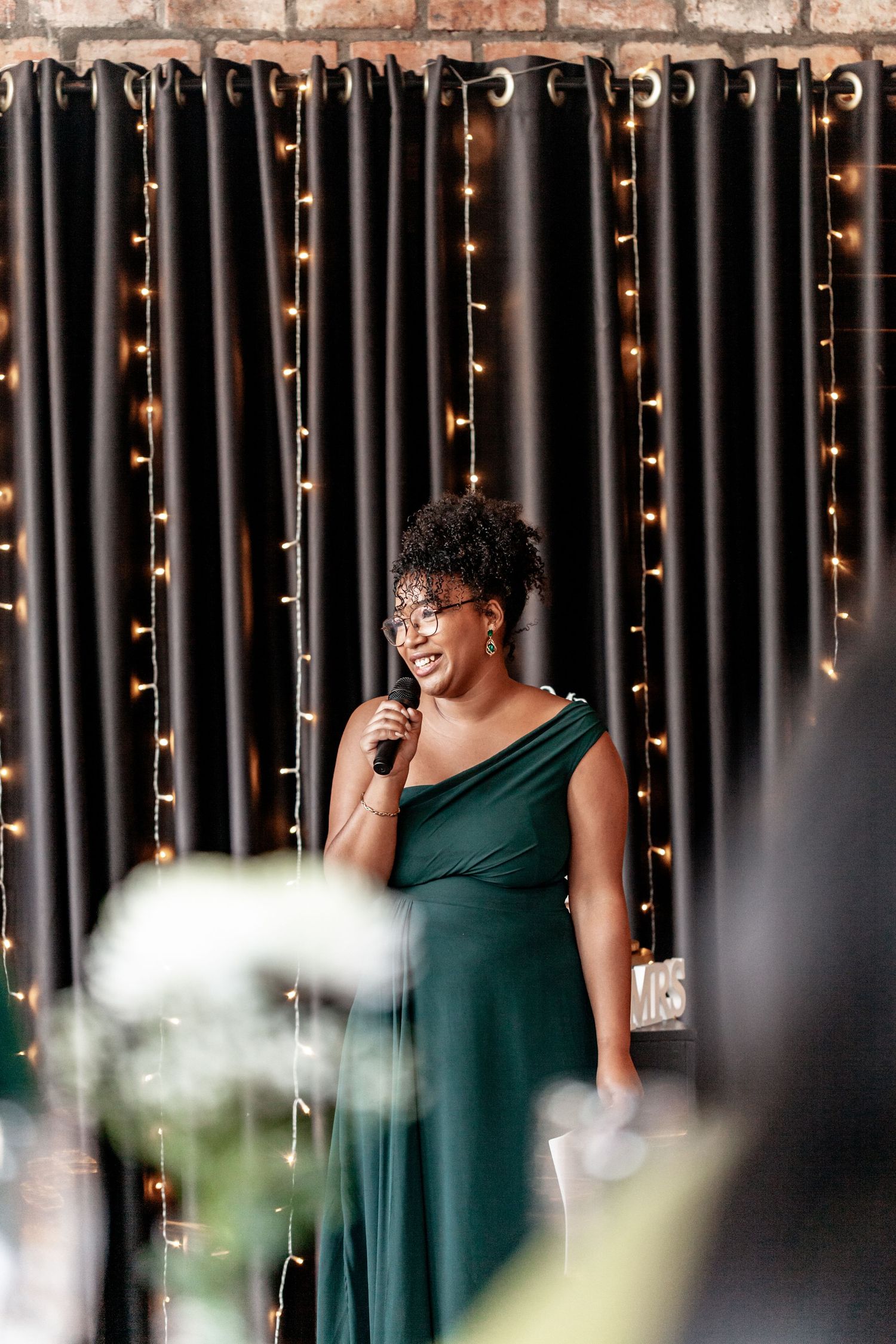 A person in an elegant green formal dress speaking into a microphone against a backdrop of twinkling string lights.