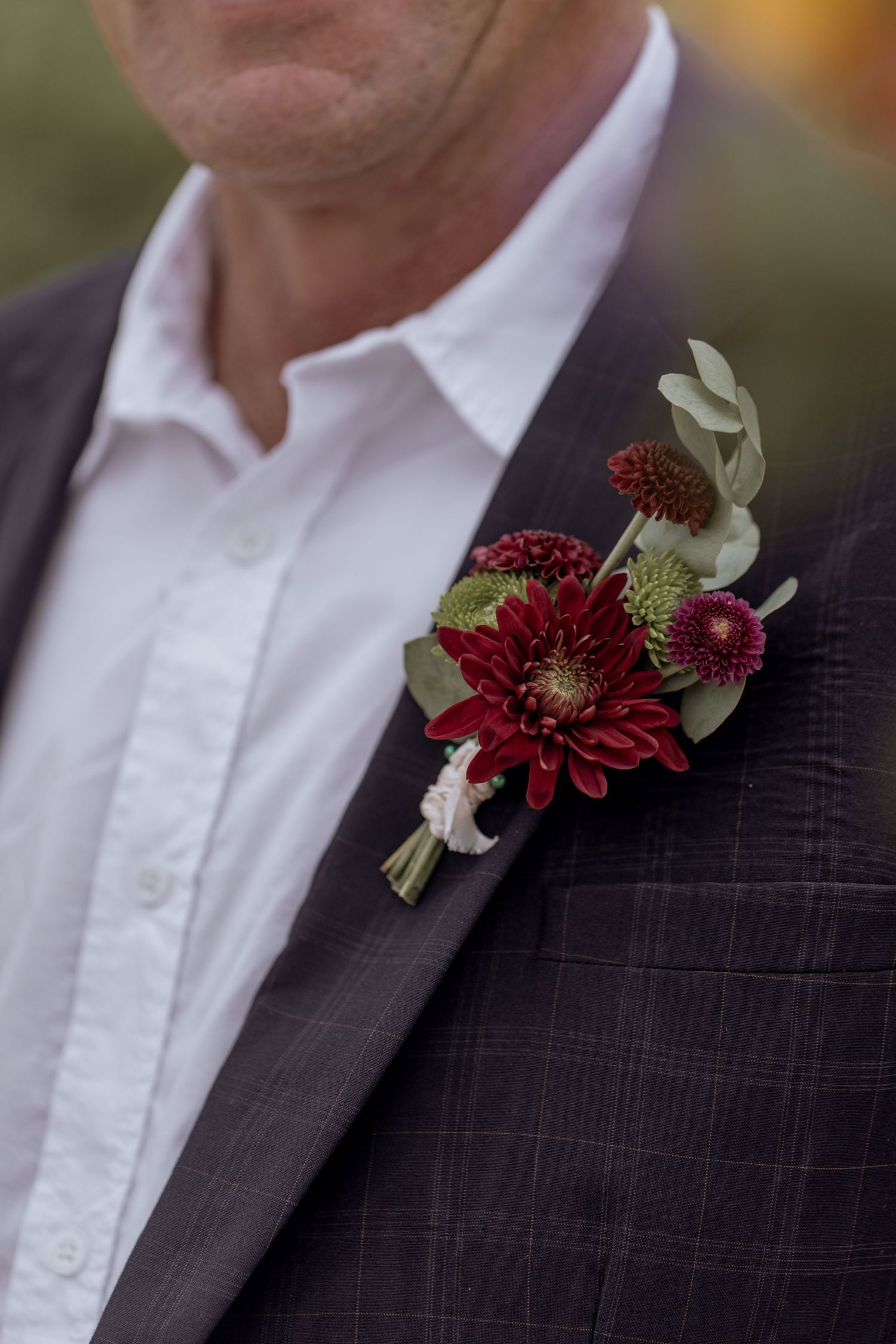 Deep burgundy floral boutonniere with delicate green ribbon adorns a dark pinstriped suit lapel at a formal event.