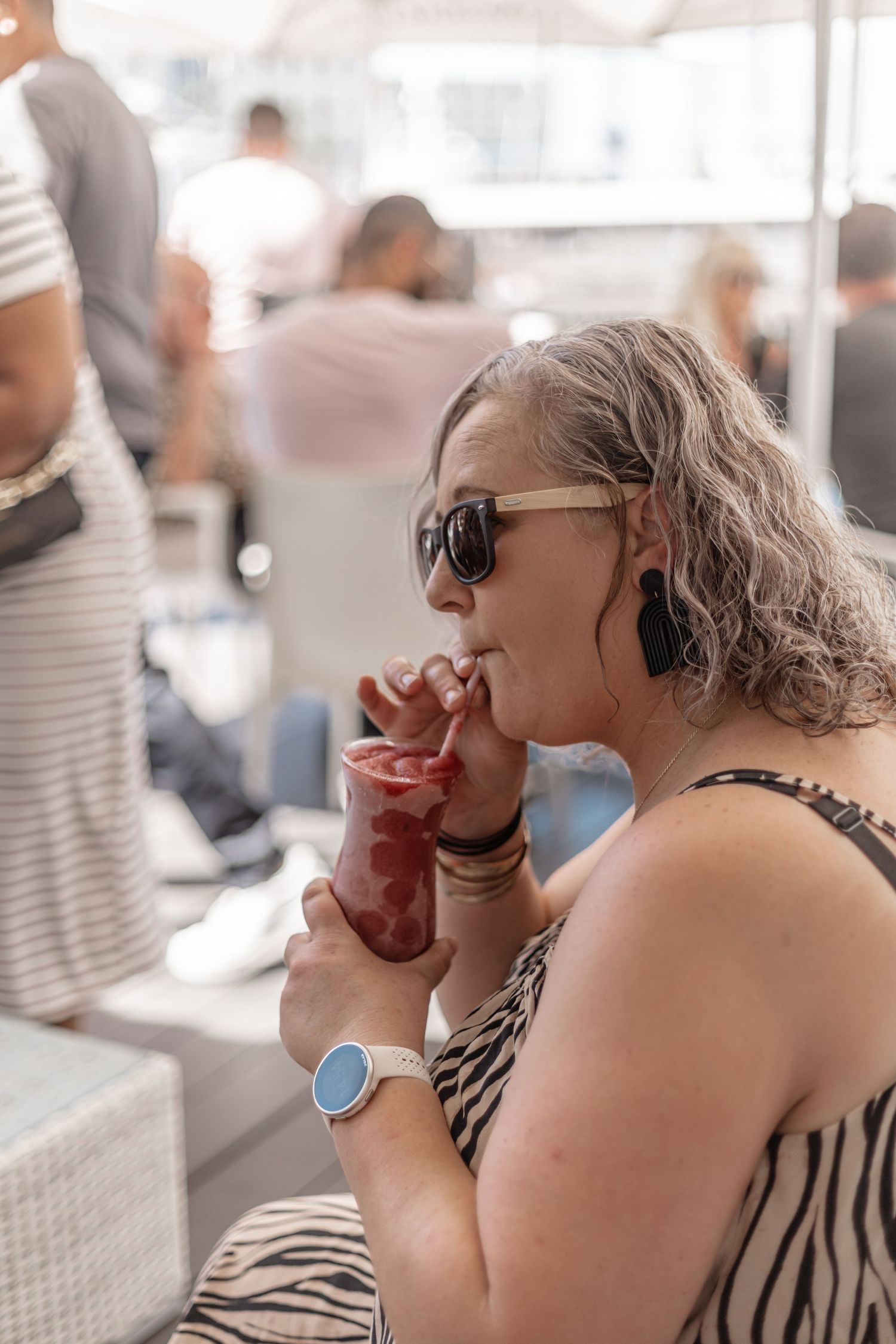 Woman with silver curly hair and sunglasses enjoying a drink at an outdoor gathering.