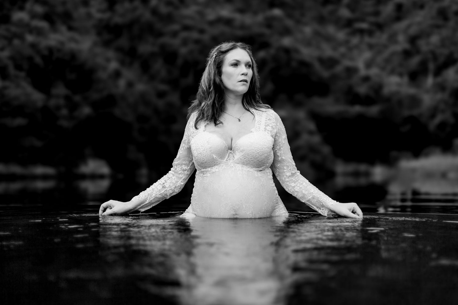 A black and white photo captures a woman wearing a long sleeve dress standing waist-deep in tranquil water.
