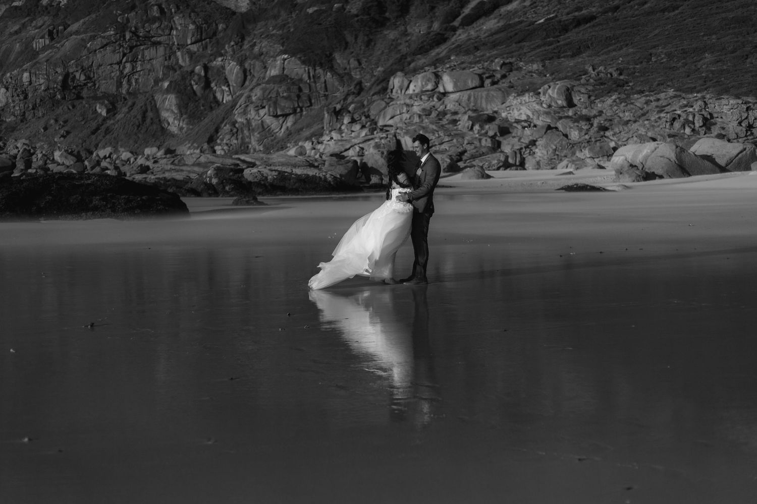 Black and white photo of a couple in wedding attire embracing on a reflective beach with rocky cliffs behind them.