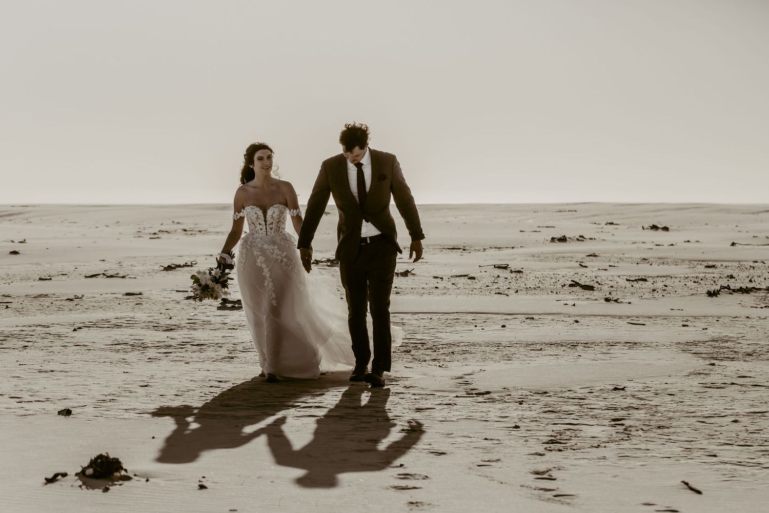 Bride and groom walk hand in hand on a sandy beach, casting long shadows in warm golden light.