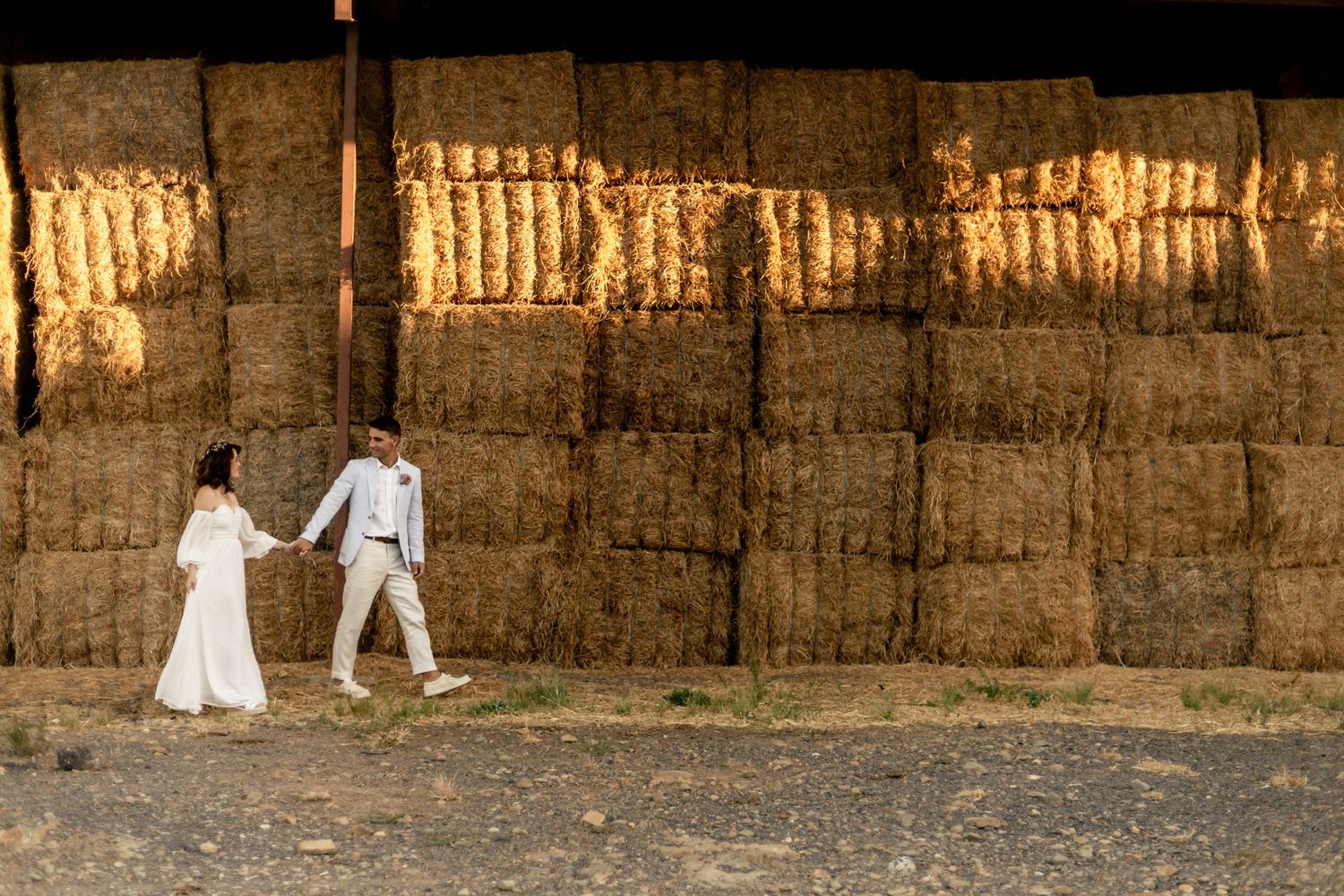 Bride and groom walk hand in hand past golden hay bales in a rustic barn at sunset.