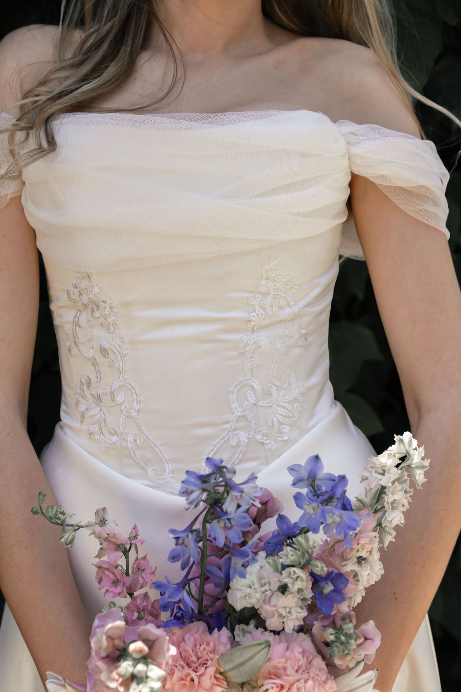A wedding dress bodice with off-shoulder sleeves and delicate floral embroidery is adorned with pink and purple flowers.