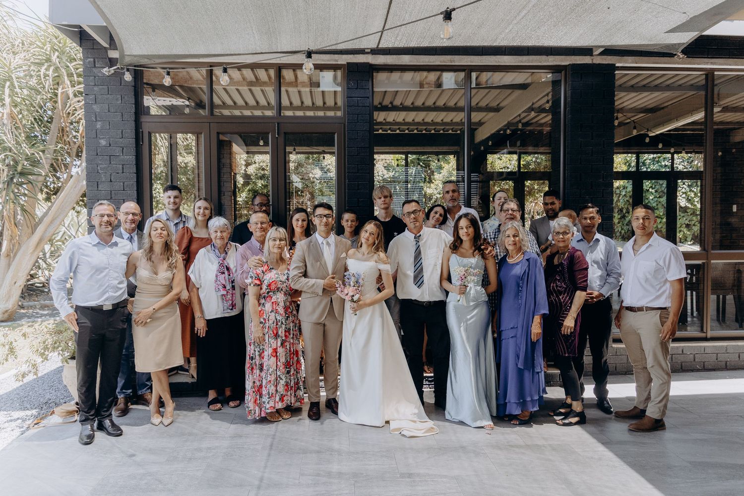 Wedding party members in formal attire gather for group photos at an outdoor venue with rustic wooden structures.