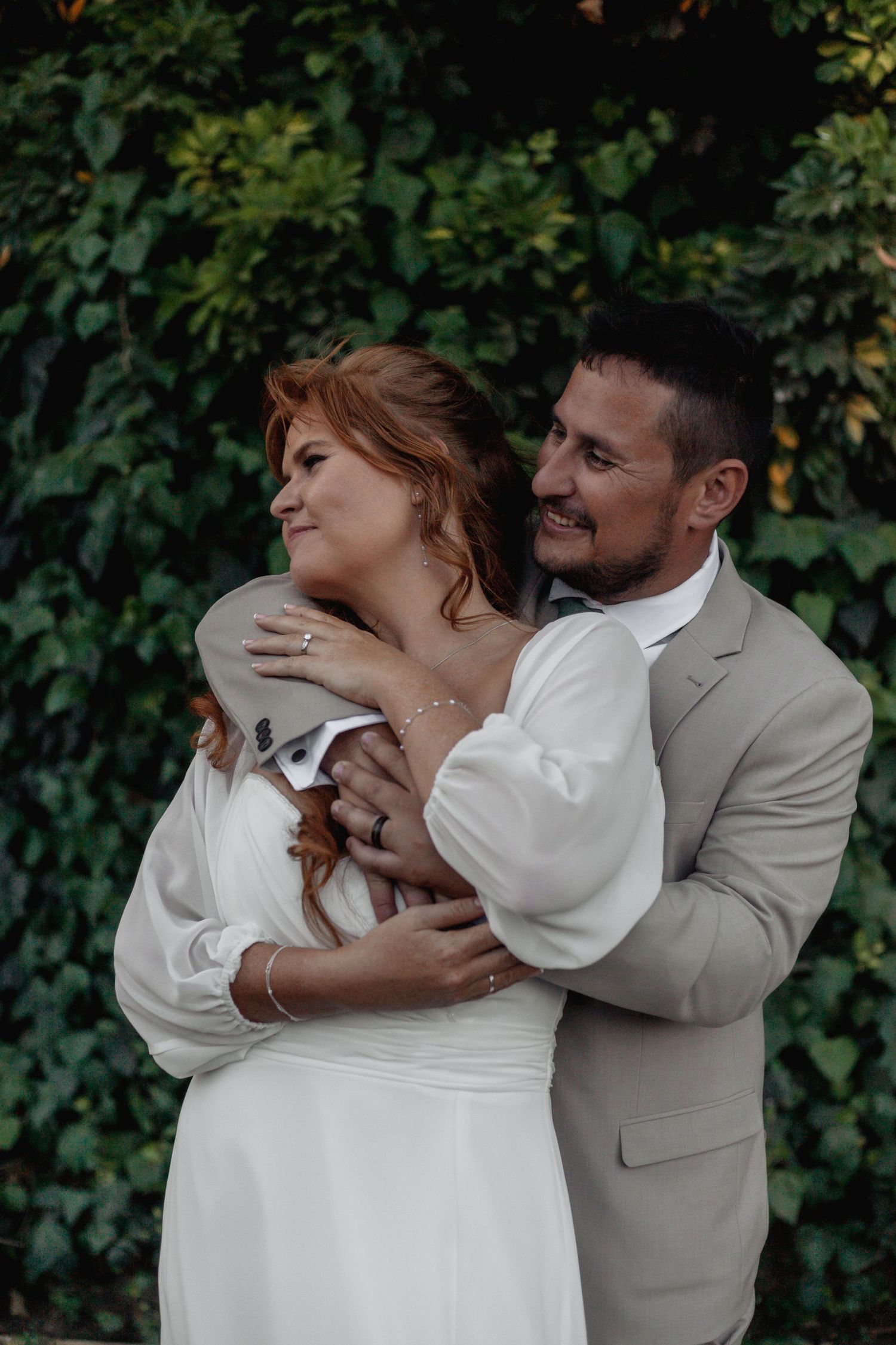 A groom in a beige suit embraces his redhead bride in a white dress, both smiling, with lush green foliage behind them.