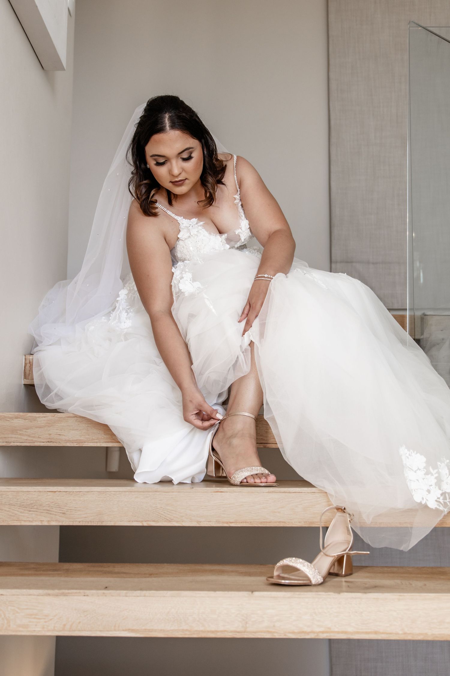 A bride in a white lace gown sits on wooden stairs while fastening her strappy heeled sandals.