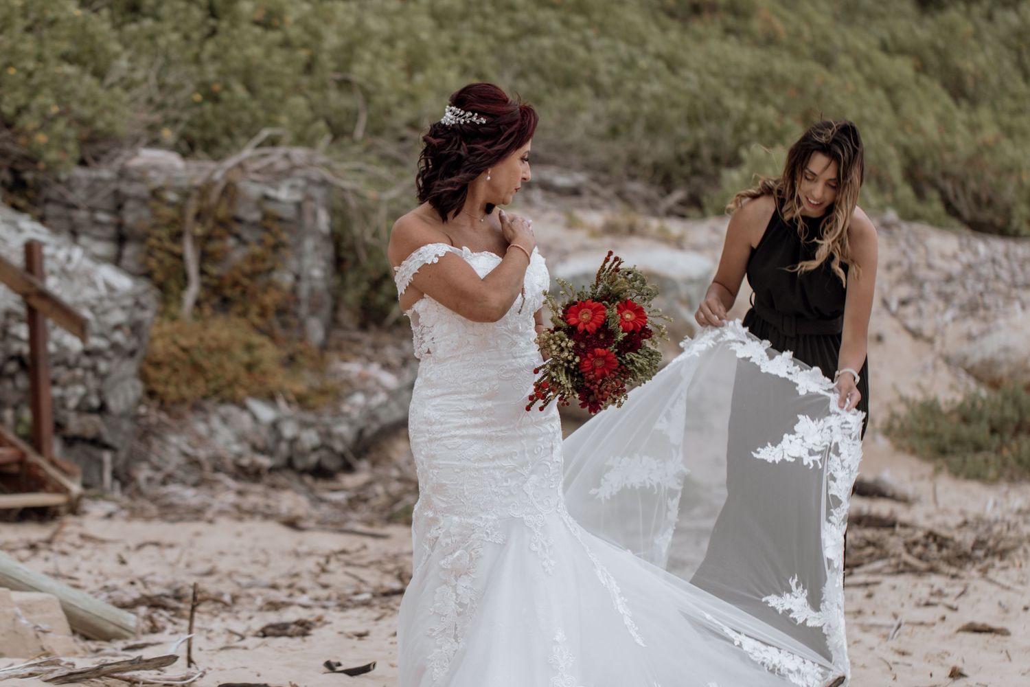A couple in wedding attire walks along a scenic rocky beach path holding hands and carrying a red bouquet.