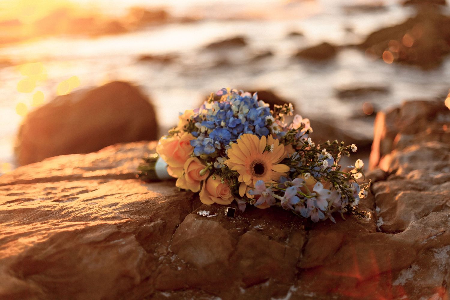 Bouquet of daisies and blue flowers resting on coastal rocks during a golden sunset.