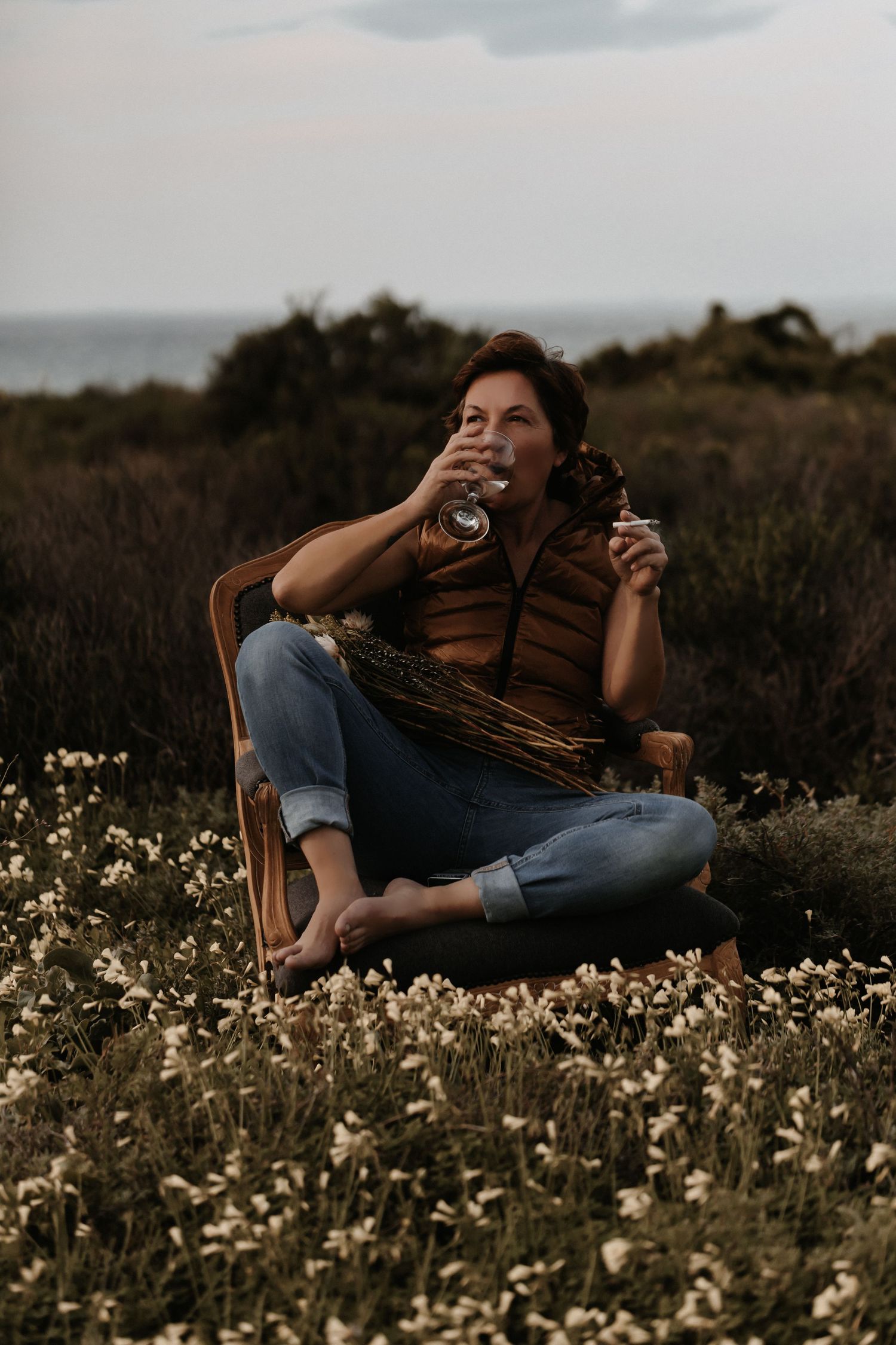 Person sitting in brown chair outdoors among wildflowers at sunset wearing jeans and striped shirt against coastal backdrop.