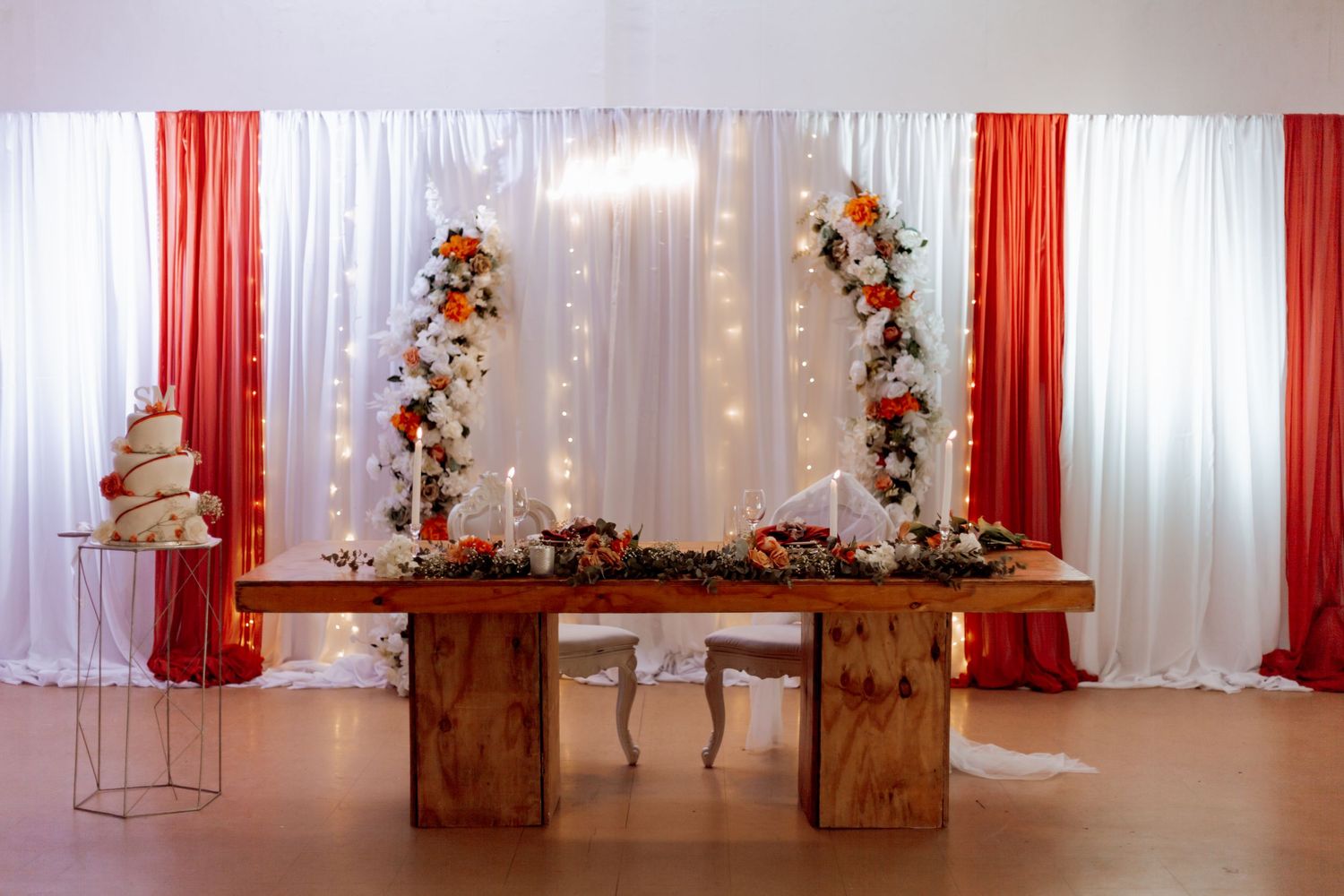 White chiffon backdrop with orange accents and rustic wooden posts creates an elegant wedding ceremony setting with twinkle lights.