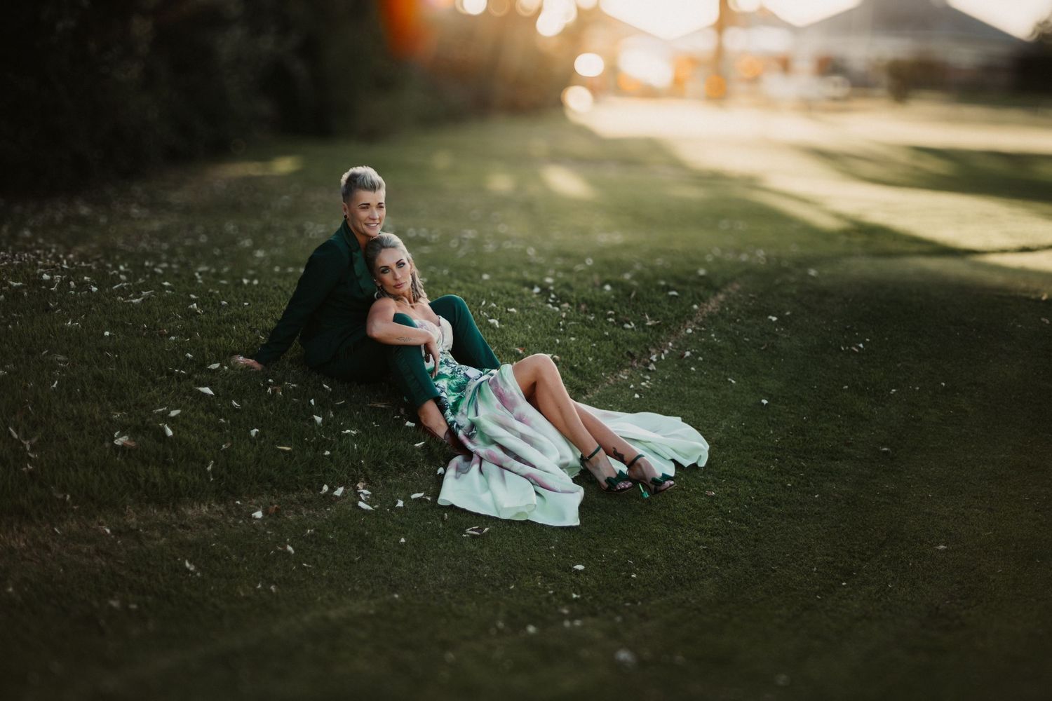 A couple sits together on a blanket in a park during golden hour as sunlight filters through the trees.