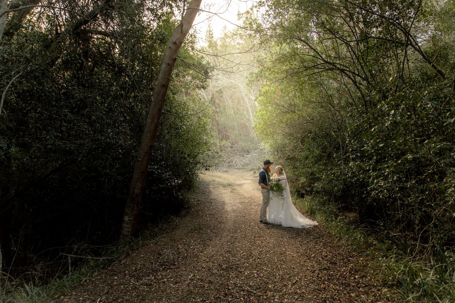 Bride and groom embracing on a sunlit forest path surrounded by lush green trees on their wedding day.