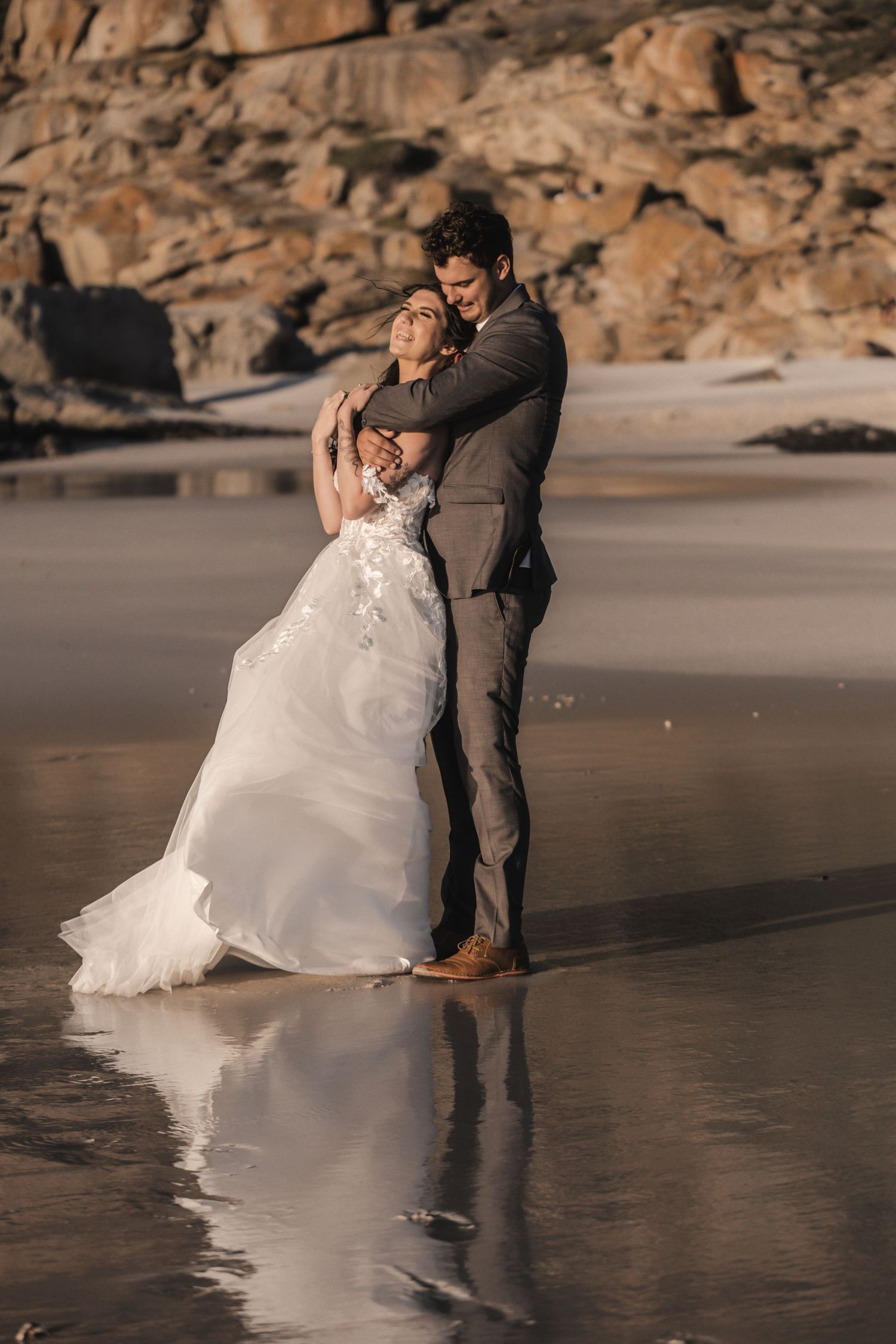 Bride and groom embrace on a reflective beach at golden hour, with rocky cliffs in the background.