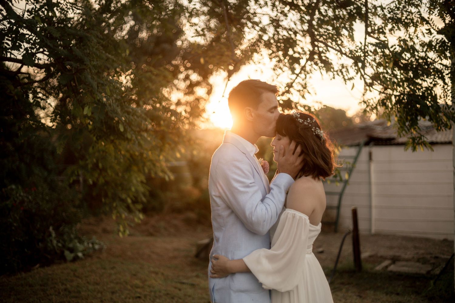 Couple sharing a romantic kiss at golden sunset in a garden, bride in white dress with floral crown.