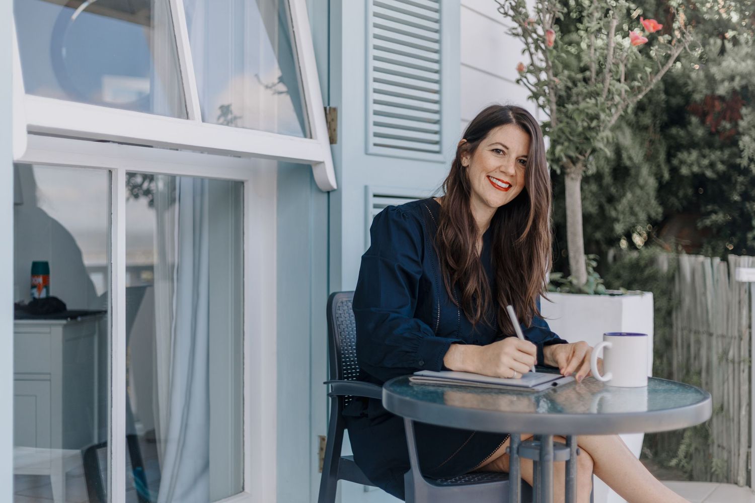 Woman in navy attire works at laptop while sitting on outdoor patio with climbing vines and floral decor.