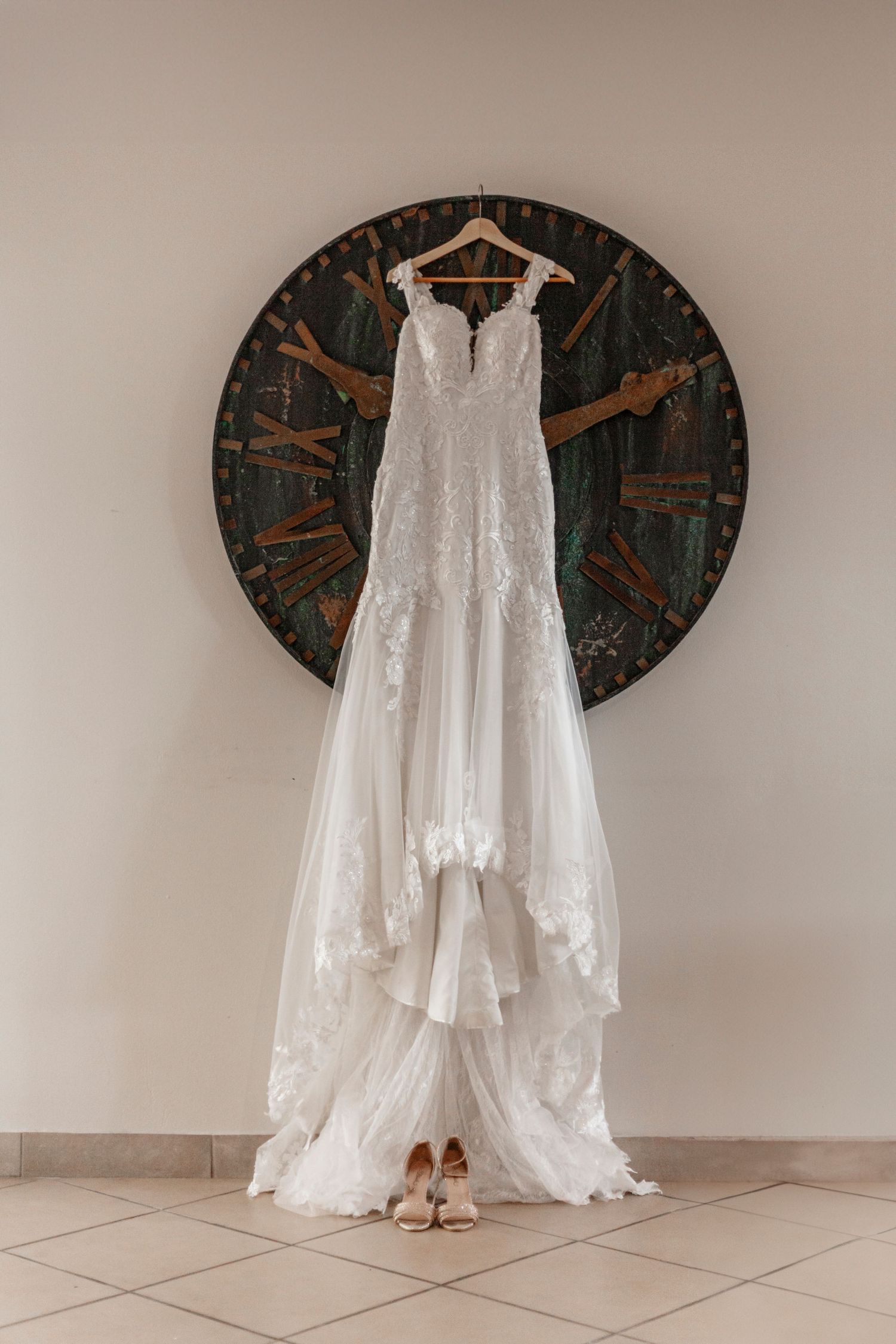 An elegant white lace wedding dress hangs on a hanger against a rustic clock backdrop with bridal shoes beneath.