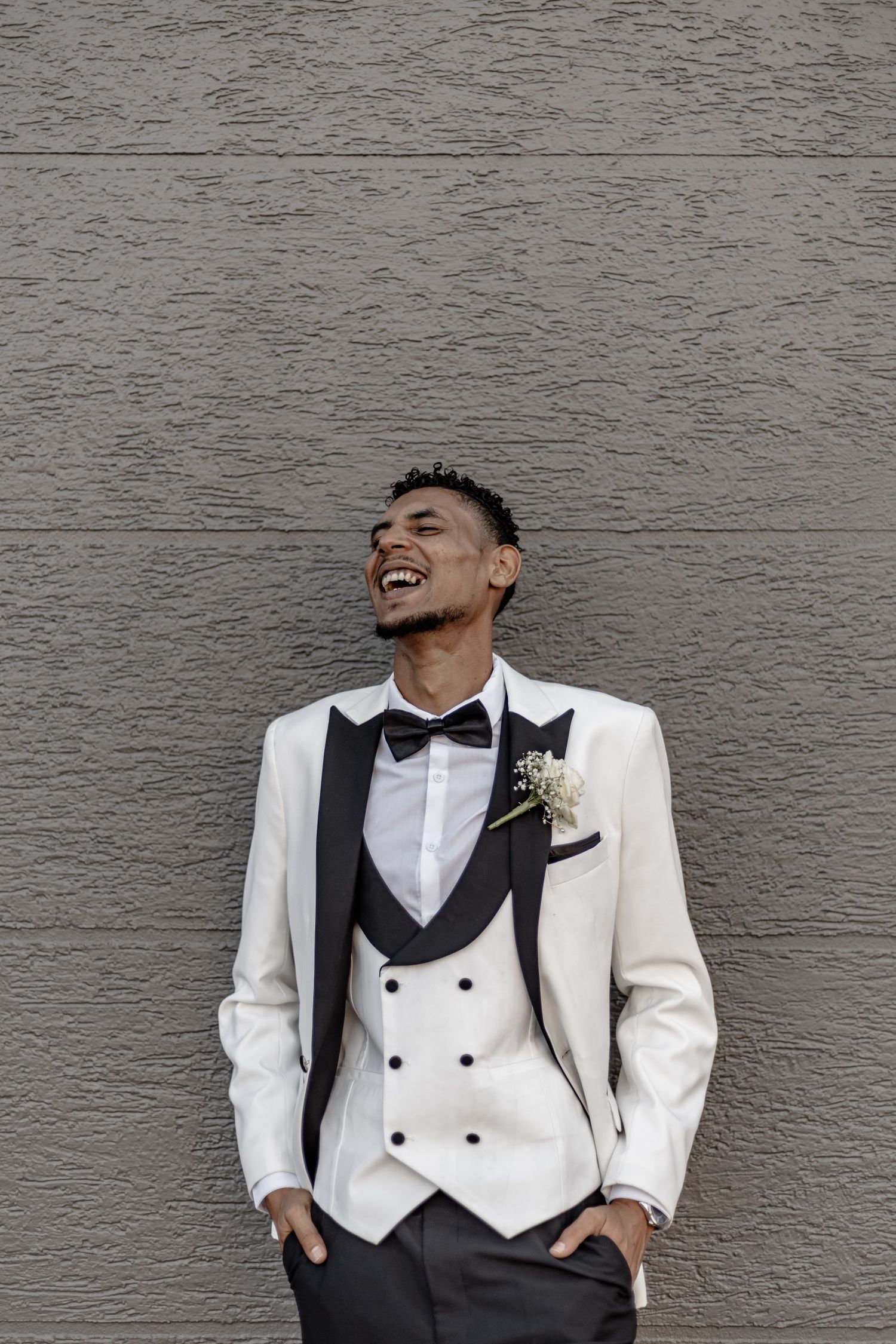 Groom in white tuxedo with black bow tie and vest stands confidently against grey wall.