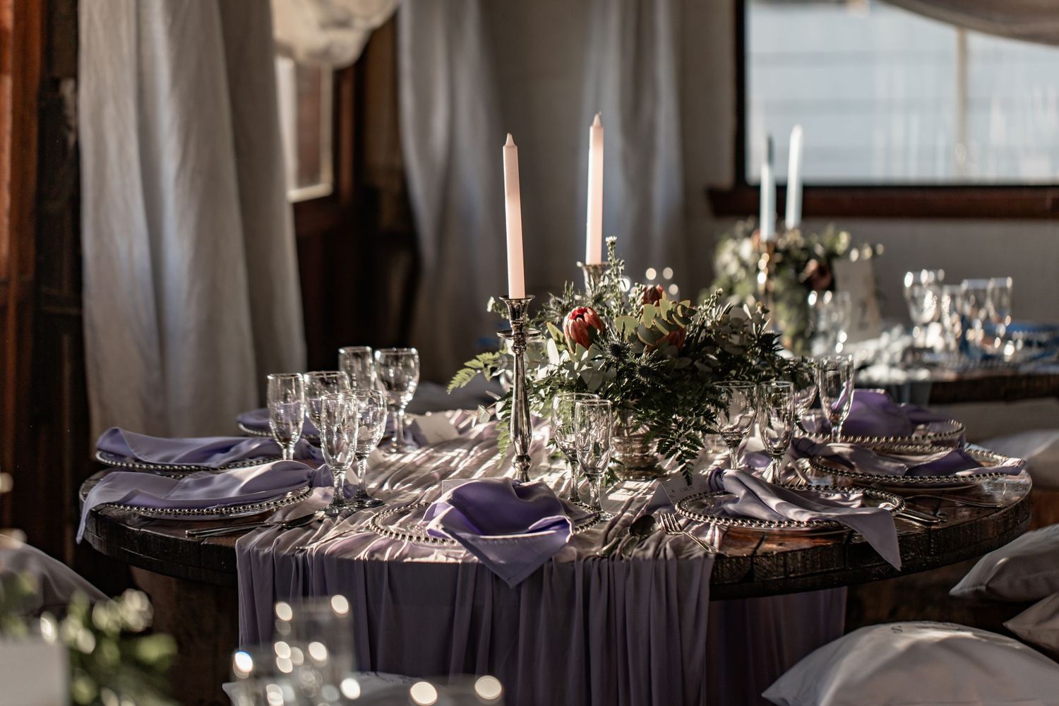 Elegant wedding table setting with tall white candles, greenery centerpiece, and dark linens in moody lighting.