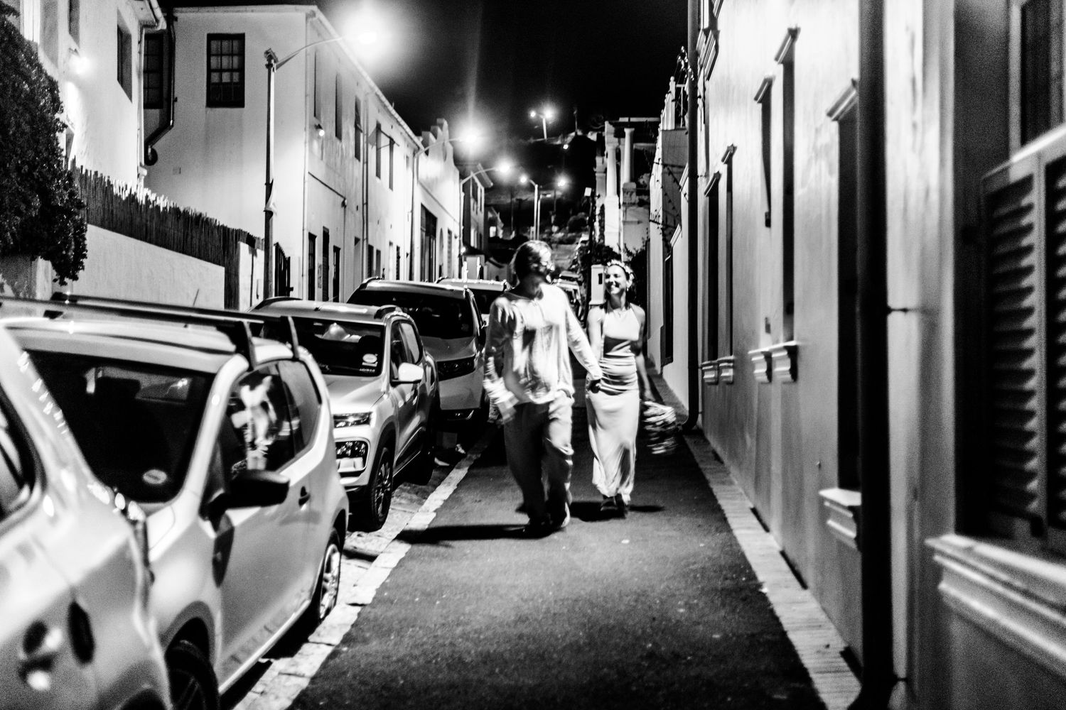 A black and white nighttime street scene shows people walking between parked cars and buildings on a narrow road.