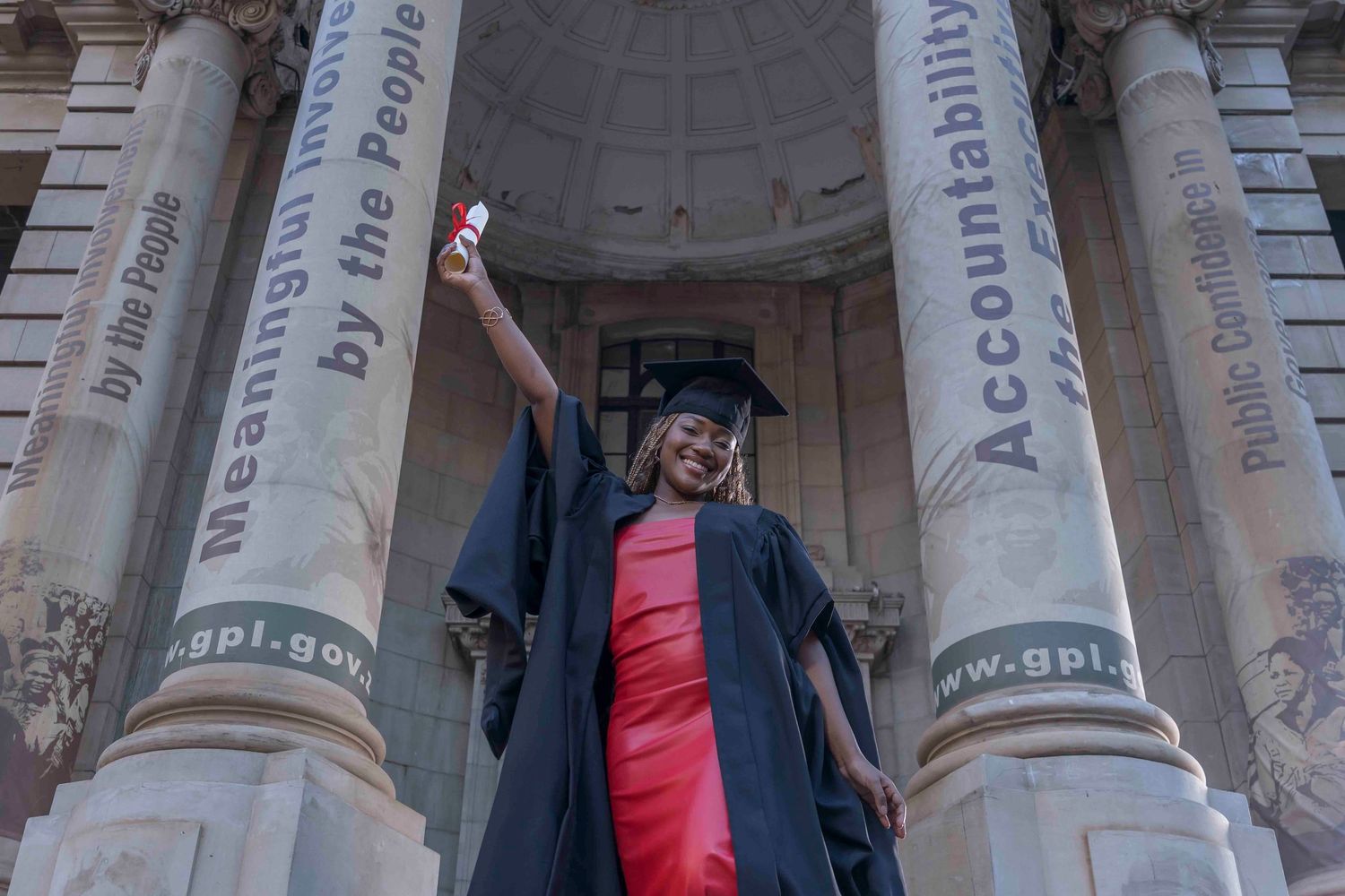 A group of graduates wearing blue caps and gowns pose for photos in front of classical stone columns.