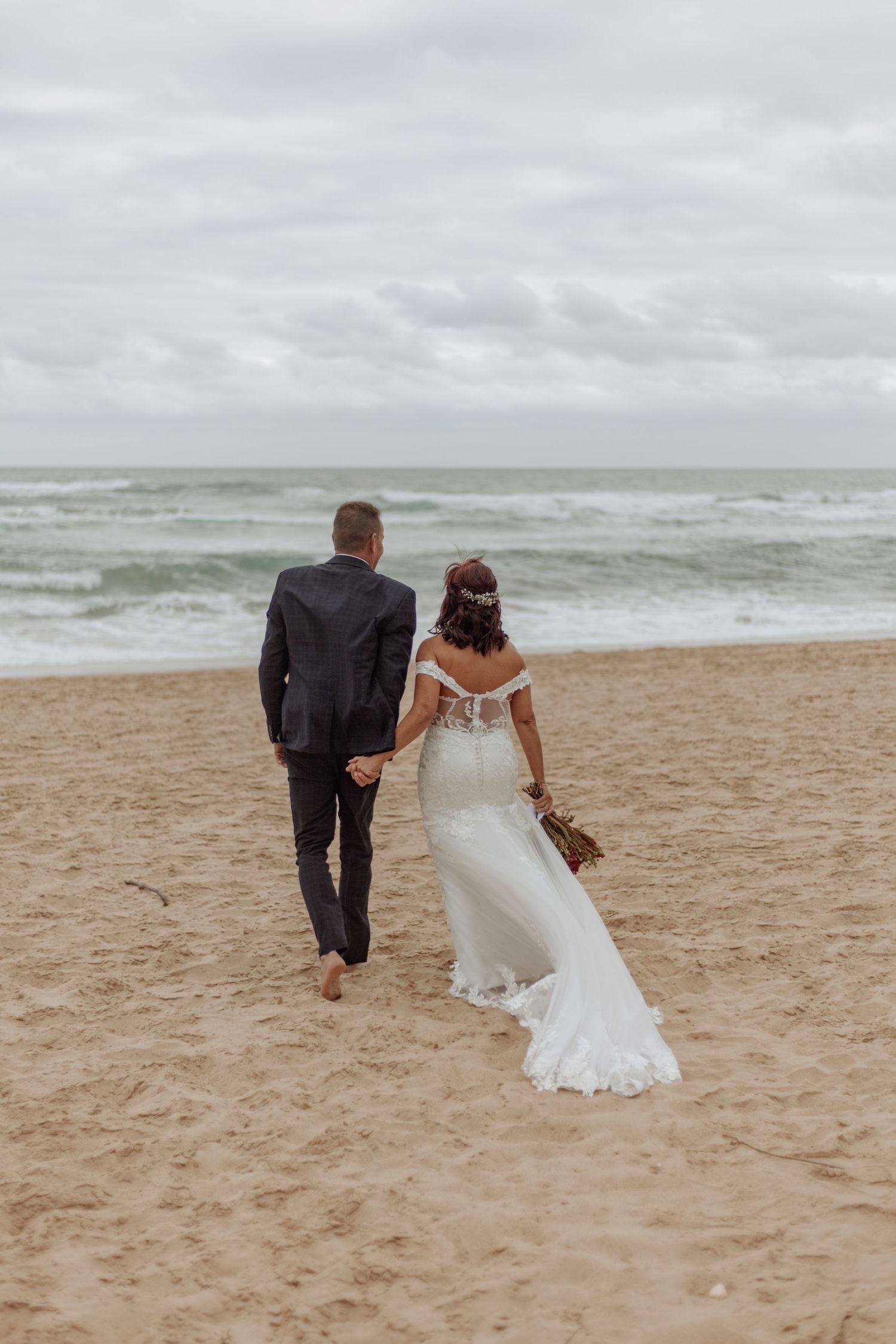 A couple walks along a moody beach shoreline, with the bride's white lace dress flowing in the ocean breeze.