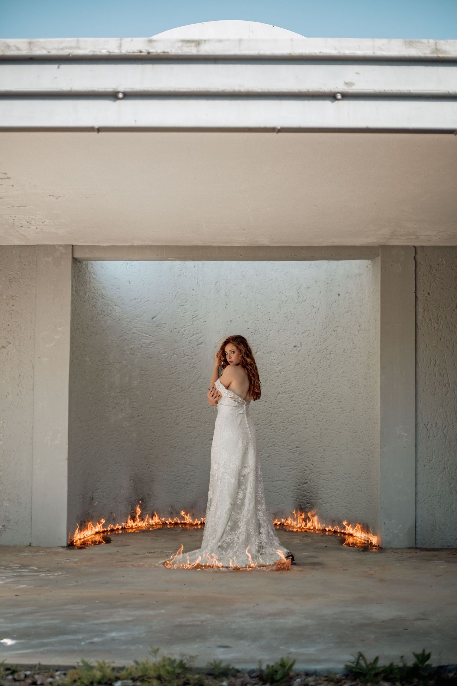 A white dress stands dramatically in a bare concrete space with flames burning in a circular pattern at its base.