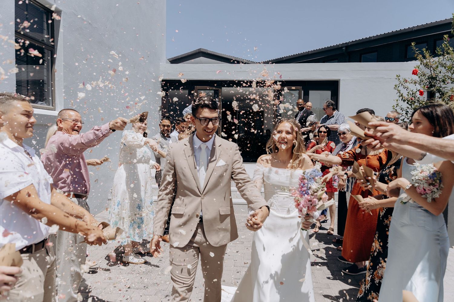 Wedding party celebrates with guests throwing confetti during outdoor celebration in beige and white attire.