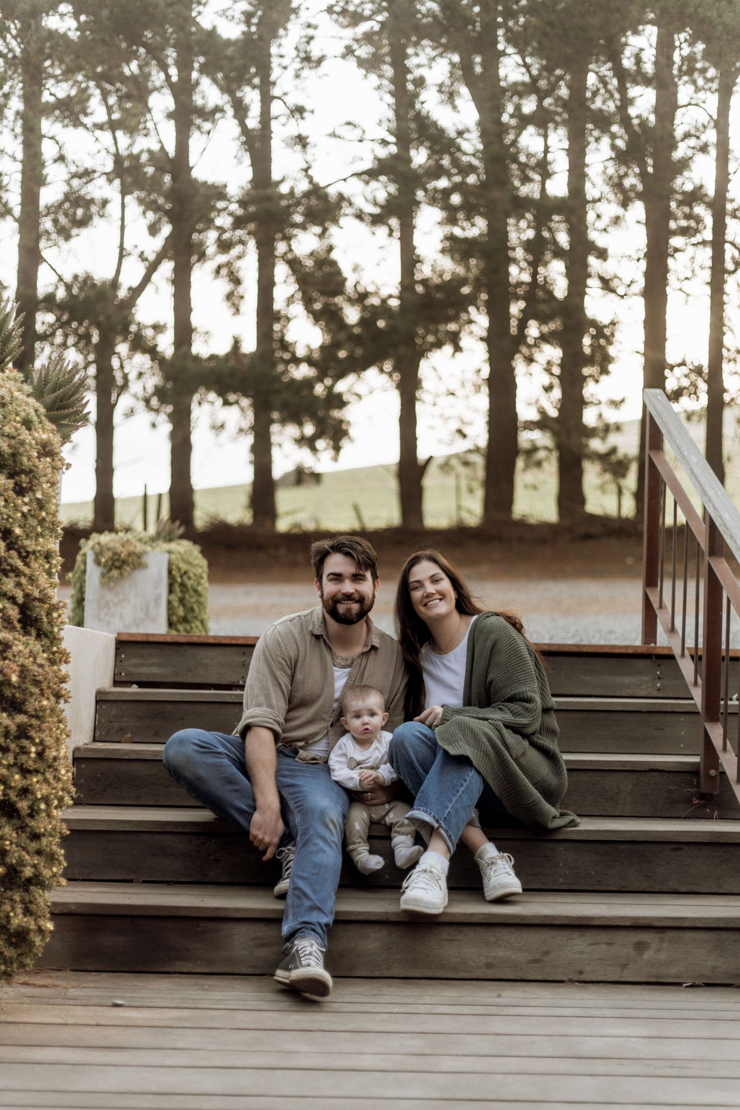 A family sits together on wooden steps outdoors surrounded by tall pine trees in a natural setting.