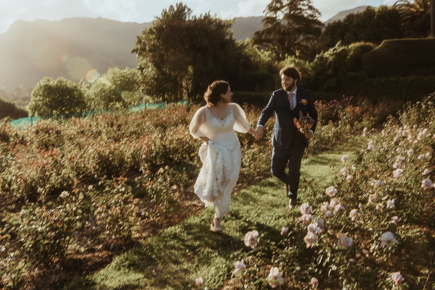 A couple walks together through a sunlit meadow filled with wildflowers during golden hour.