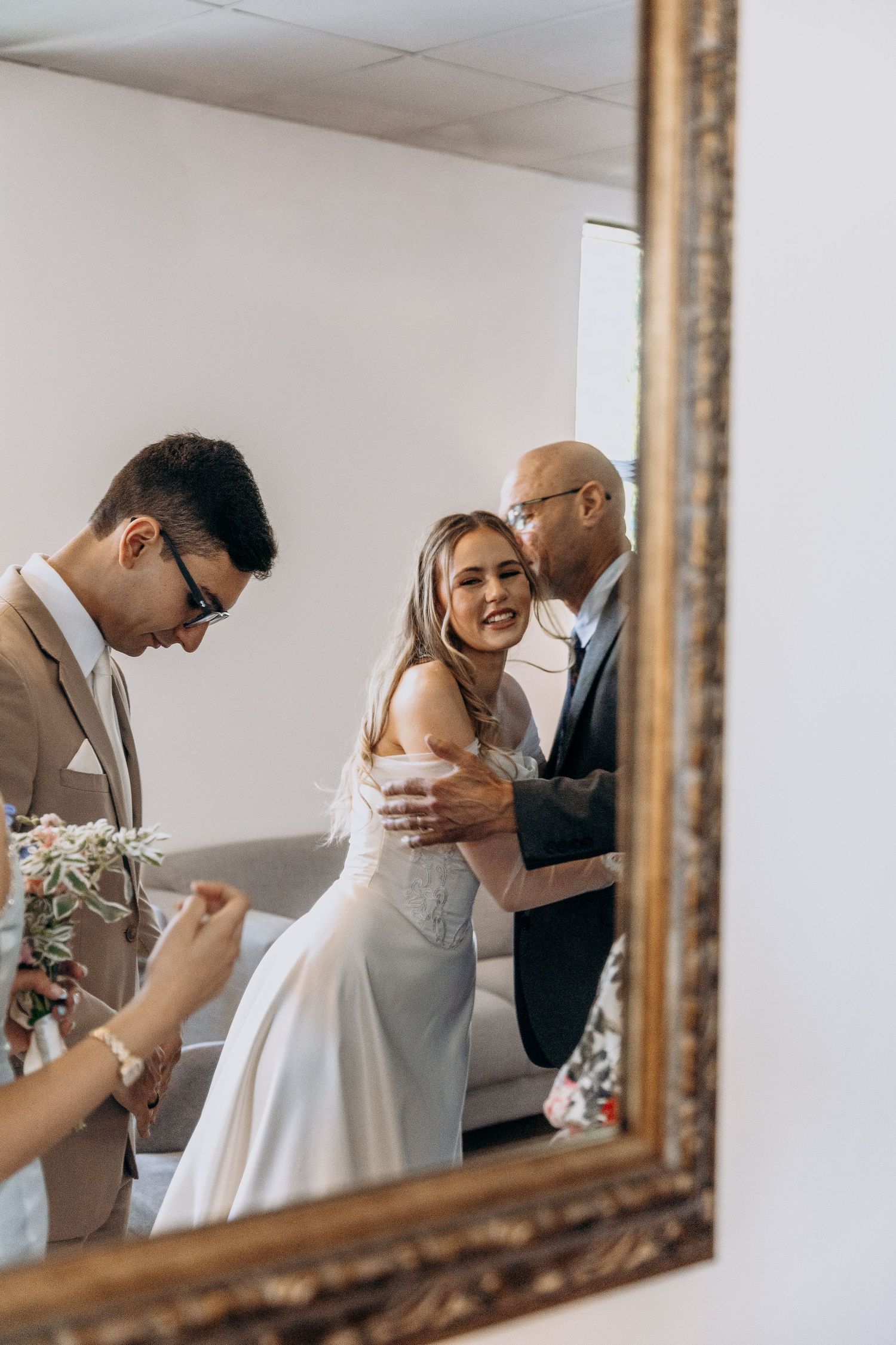 A reflection in an ornate gold mirror shows a couple sharing a tender moment while another person writes nearby.