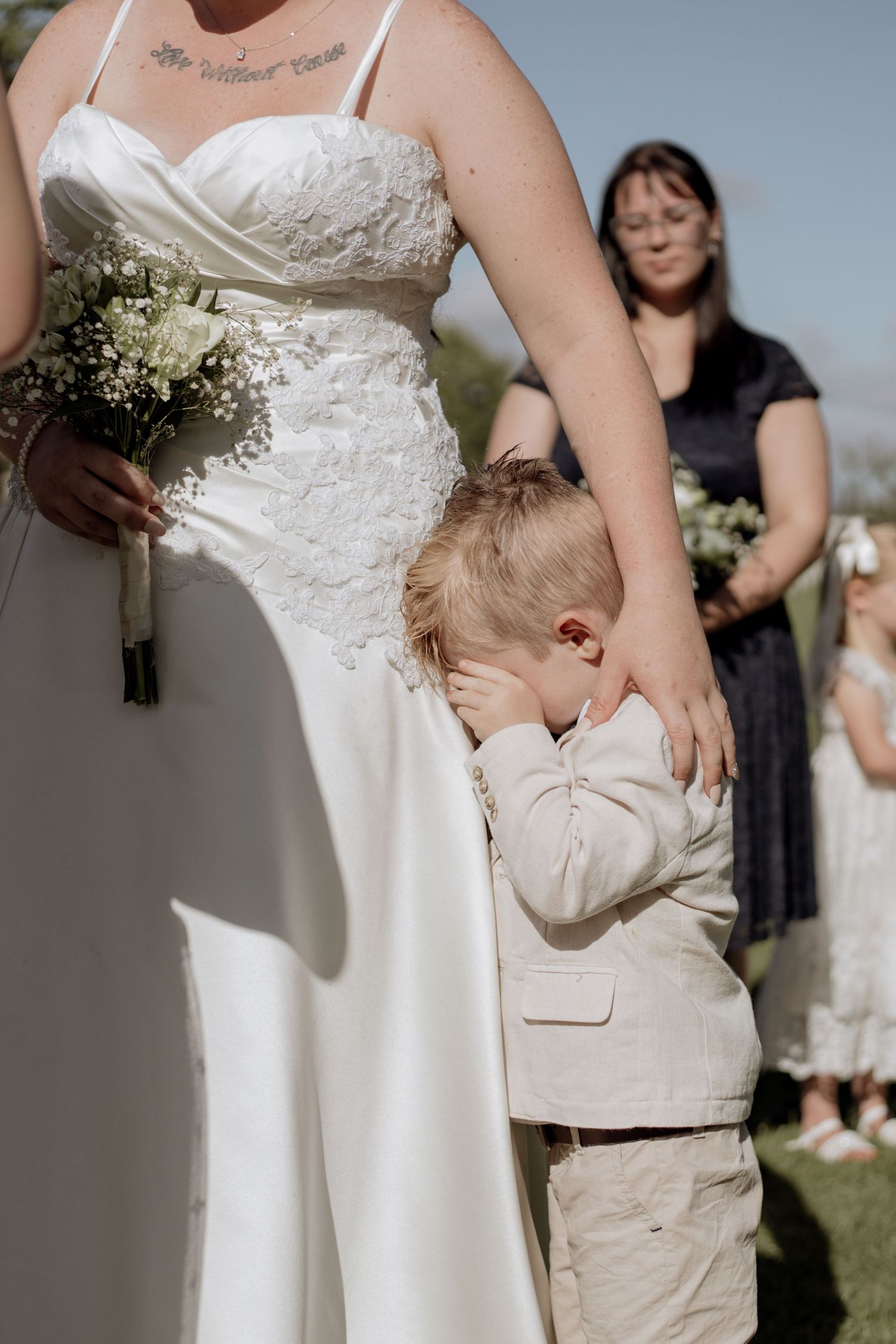 A young child in a beige suit hugs the skirt of a white wedding dress as a figure stands behind them outdoors.