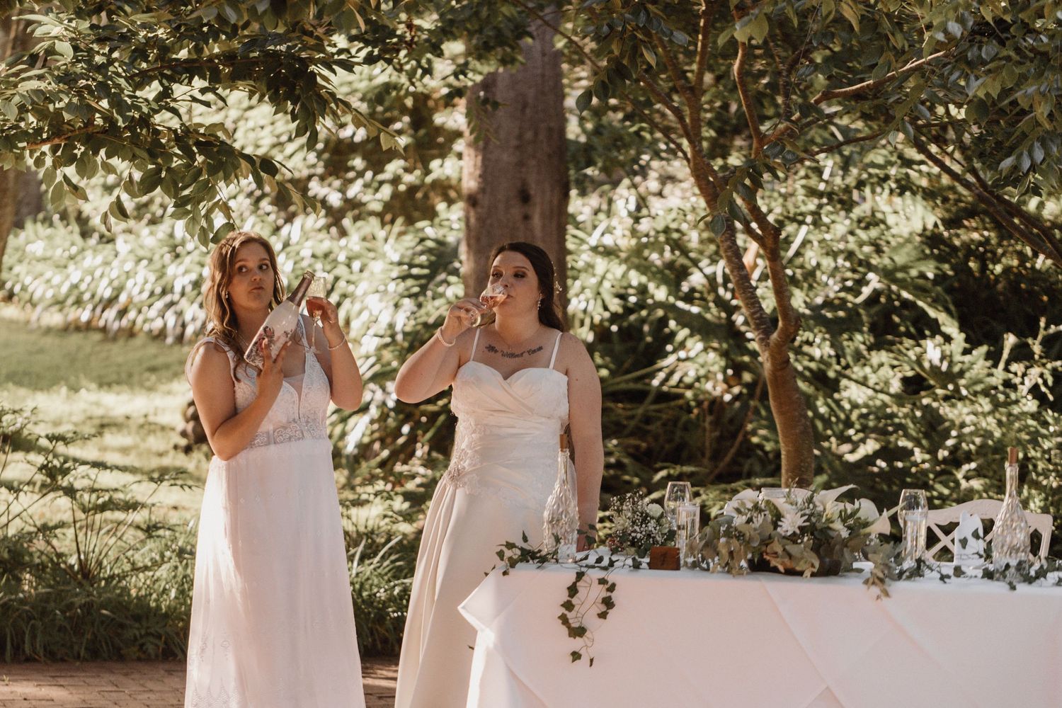 Two women in white dresses stand outdoors during a sunny wedding ceremony with floral decorations.
