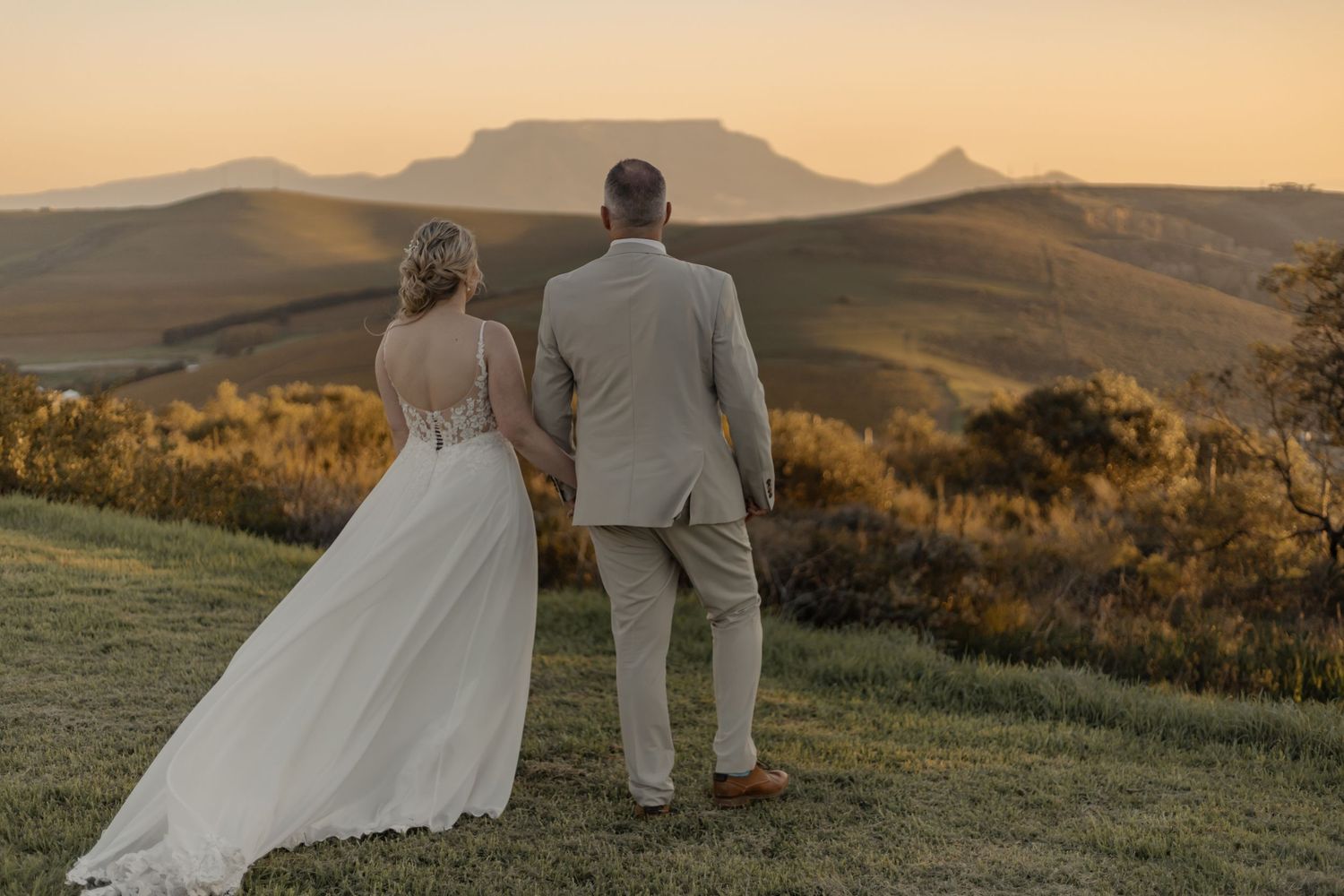 A couple in wedding attire sits together on a hillside at sunset with golden mountains in the distance.