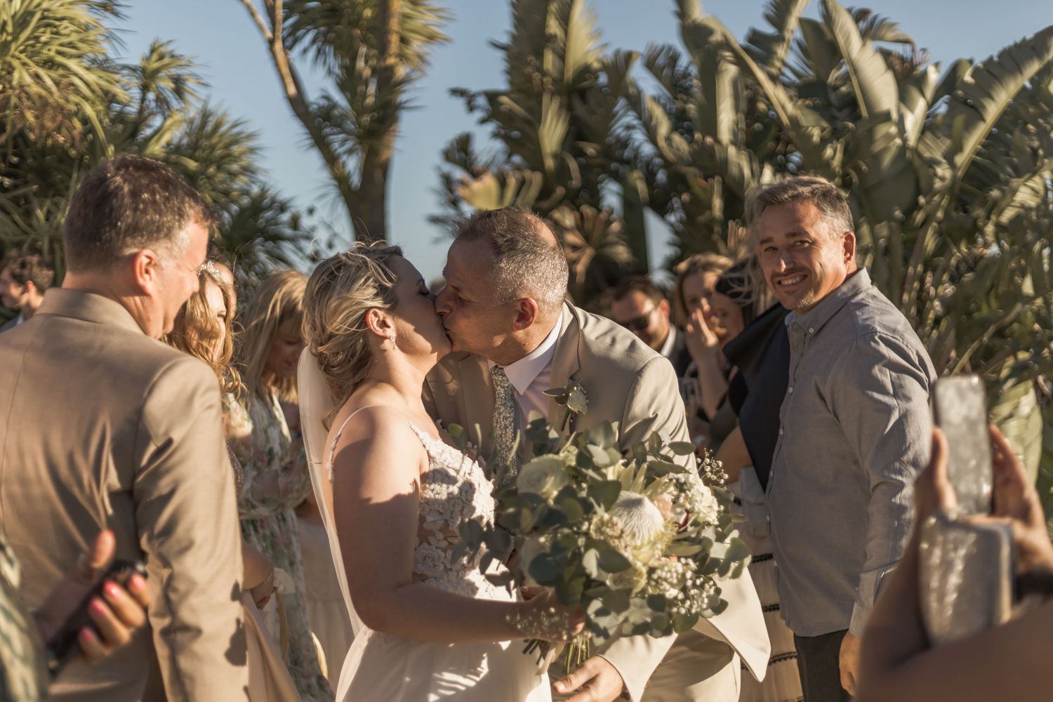 A couple shares an intimate moment during an outdoor wedding ceremony with palm trees swaying in the background at sunset.