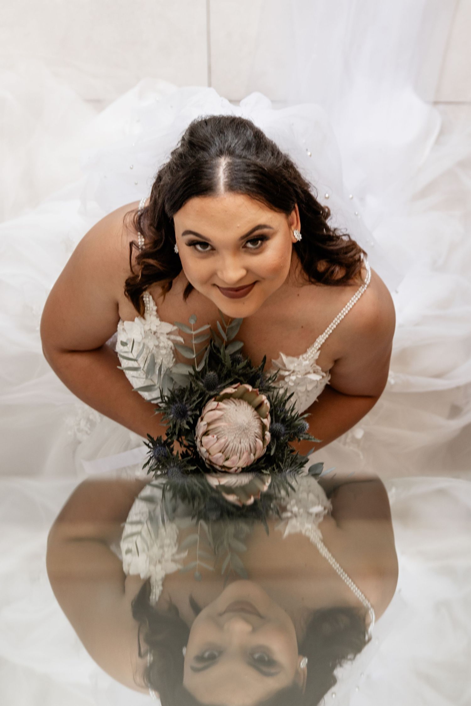 Bride in white dress leans forward with rustic bouquet and reflection on mirrored surface below.
