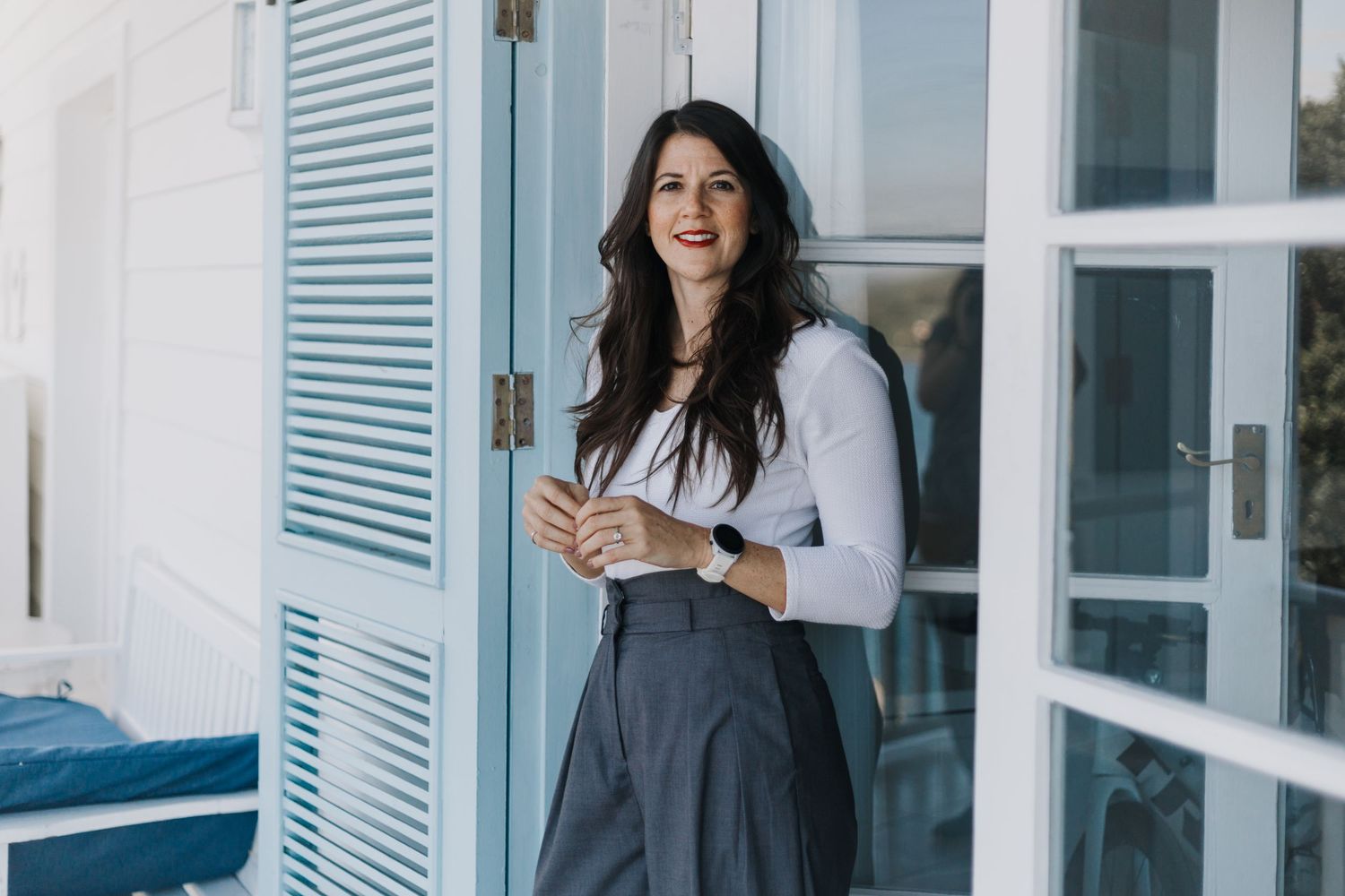 Woman with dark hair stands smiling indoors near large windows and shutters.