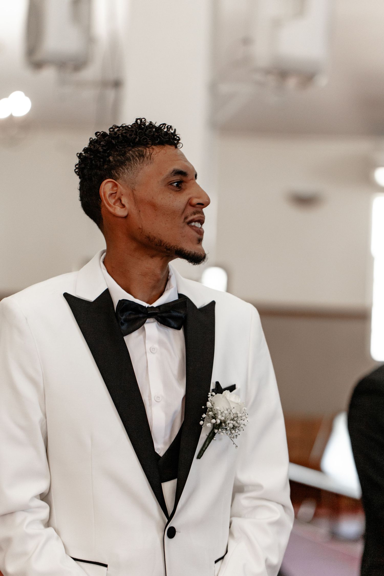 A person in a crisp white tuxedo with black lapels and bowtie smiles while looking to the side at a formal event.