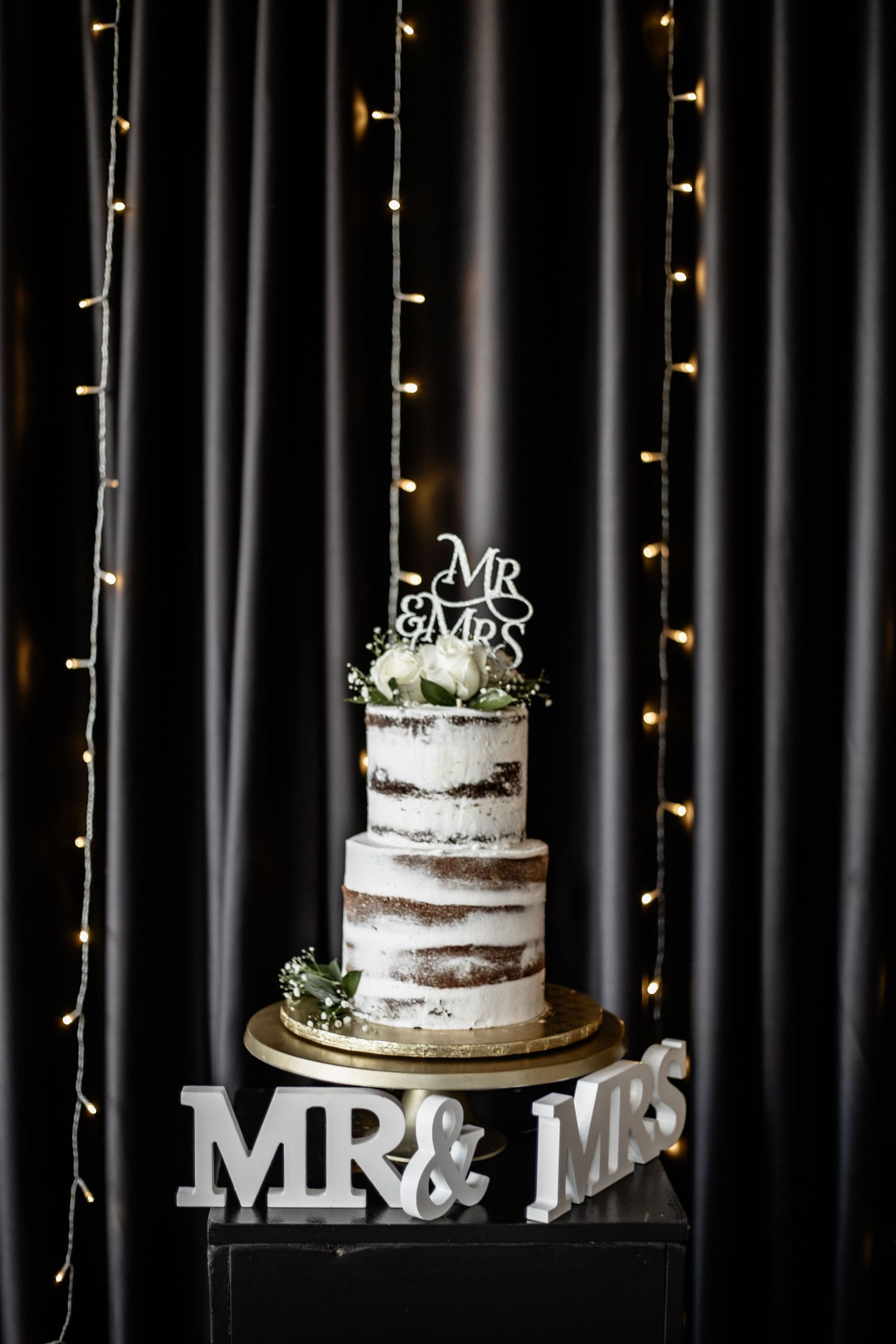 Rustic naked wedding cake with white frosting and Mr & Mrs topper displayed against black backdrop with twinkle lights.