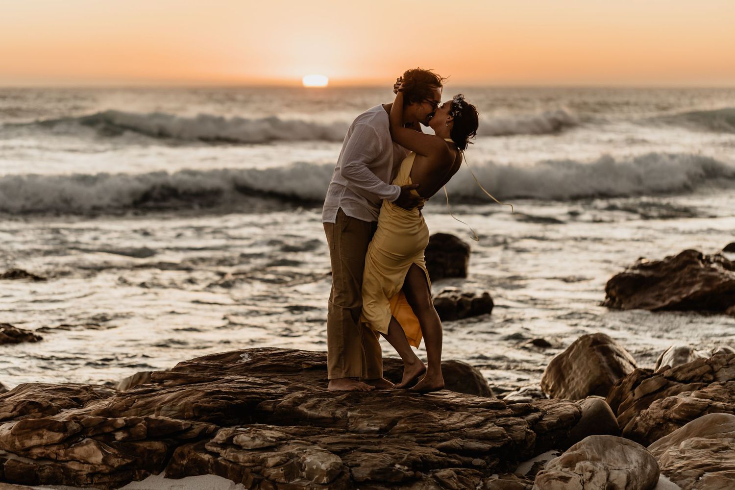 A couple shares a romantic kiss on rocky beach cliffs during golden hour sunset as waves crash behind them.