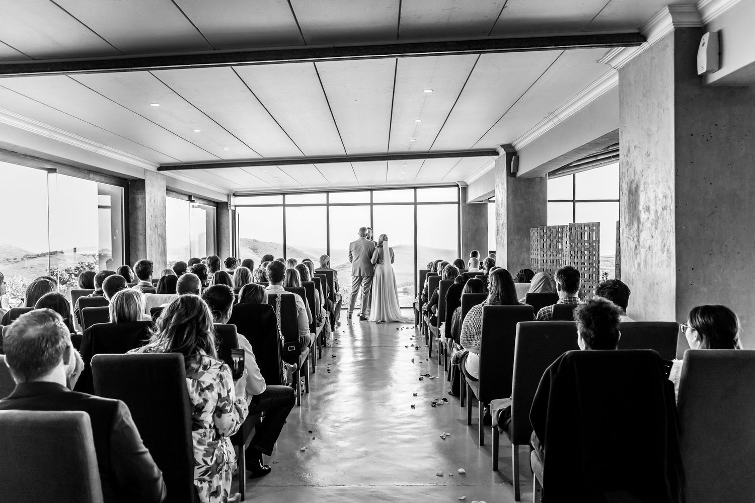 Wedding ceremony with guests sitting in rows watching a couple exchanging vows in front of large windows.