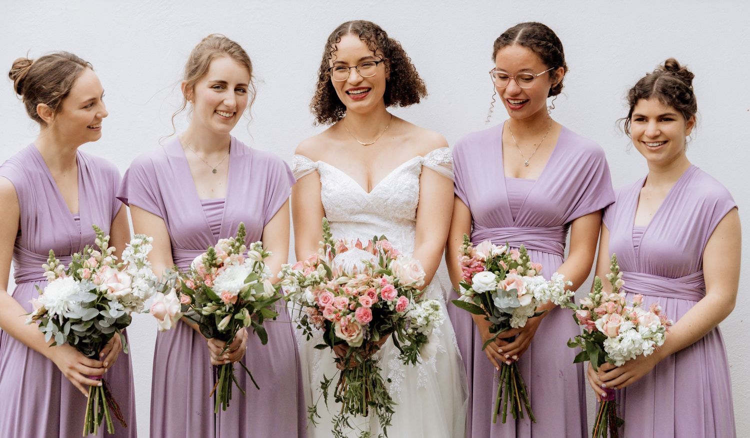 Wedding party members in matching lavender dresses hold bouquets of pink and white flowers while smiling.