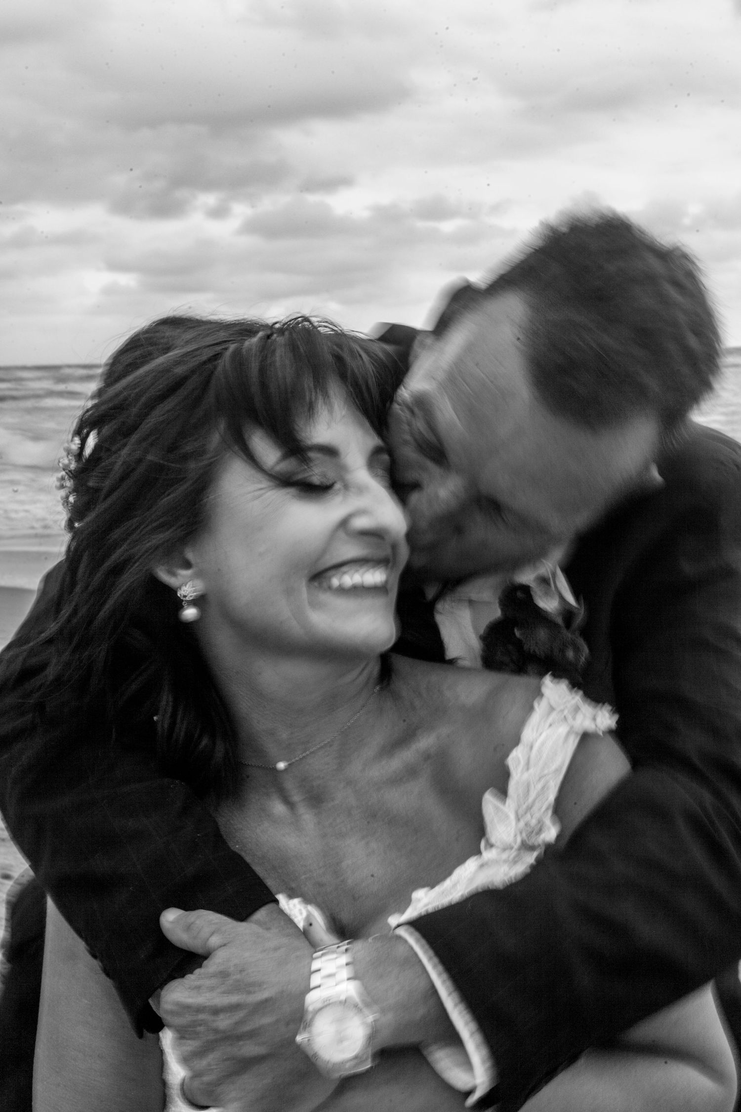 A romantic black and white photo of a couple in loving embrace at the beach with cloudy skies above.