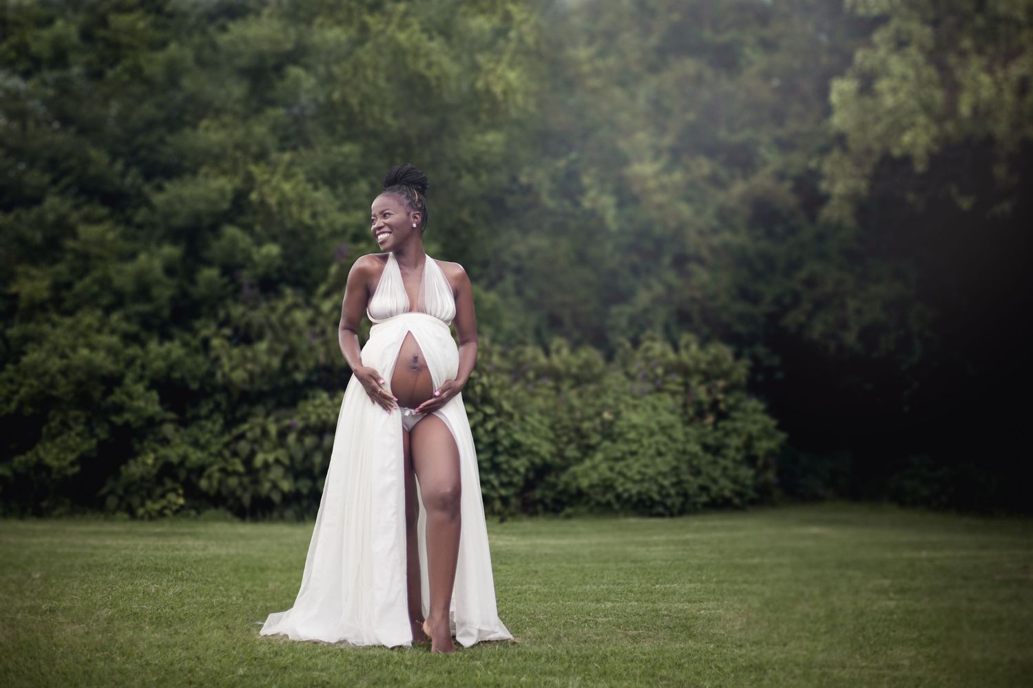 A pregnant figure stands in a flowing white gown on a grassy lawn with lush greenery in the background.