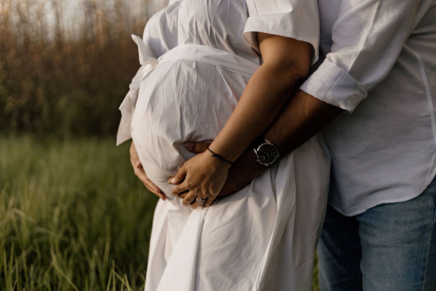 A couple in white clothing embrace in a grassy field during golden hour, with focus on their intertwined hands.