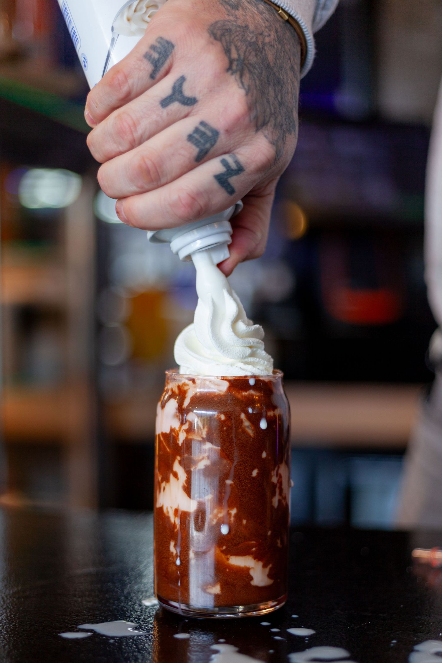 A tattooed hand pours whipped cream into a glass filled with chocolate dessert or beverage.