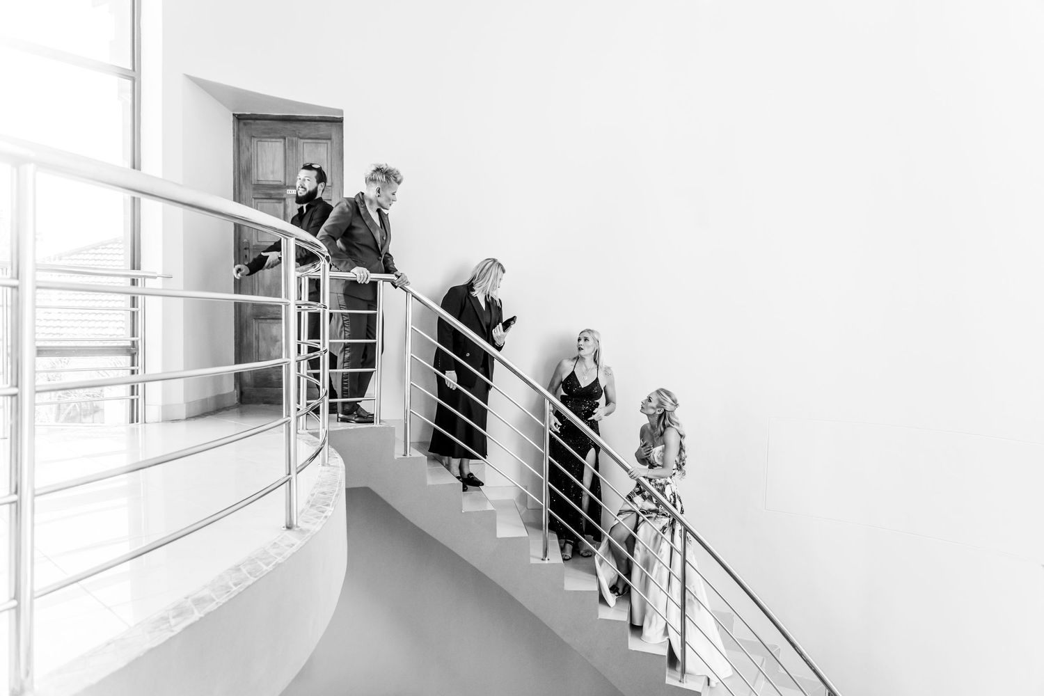 People walk along a modern white hallway with glass walls and metal railings in a minimalist architectural setting.