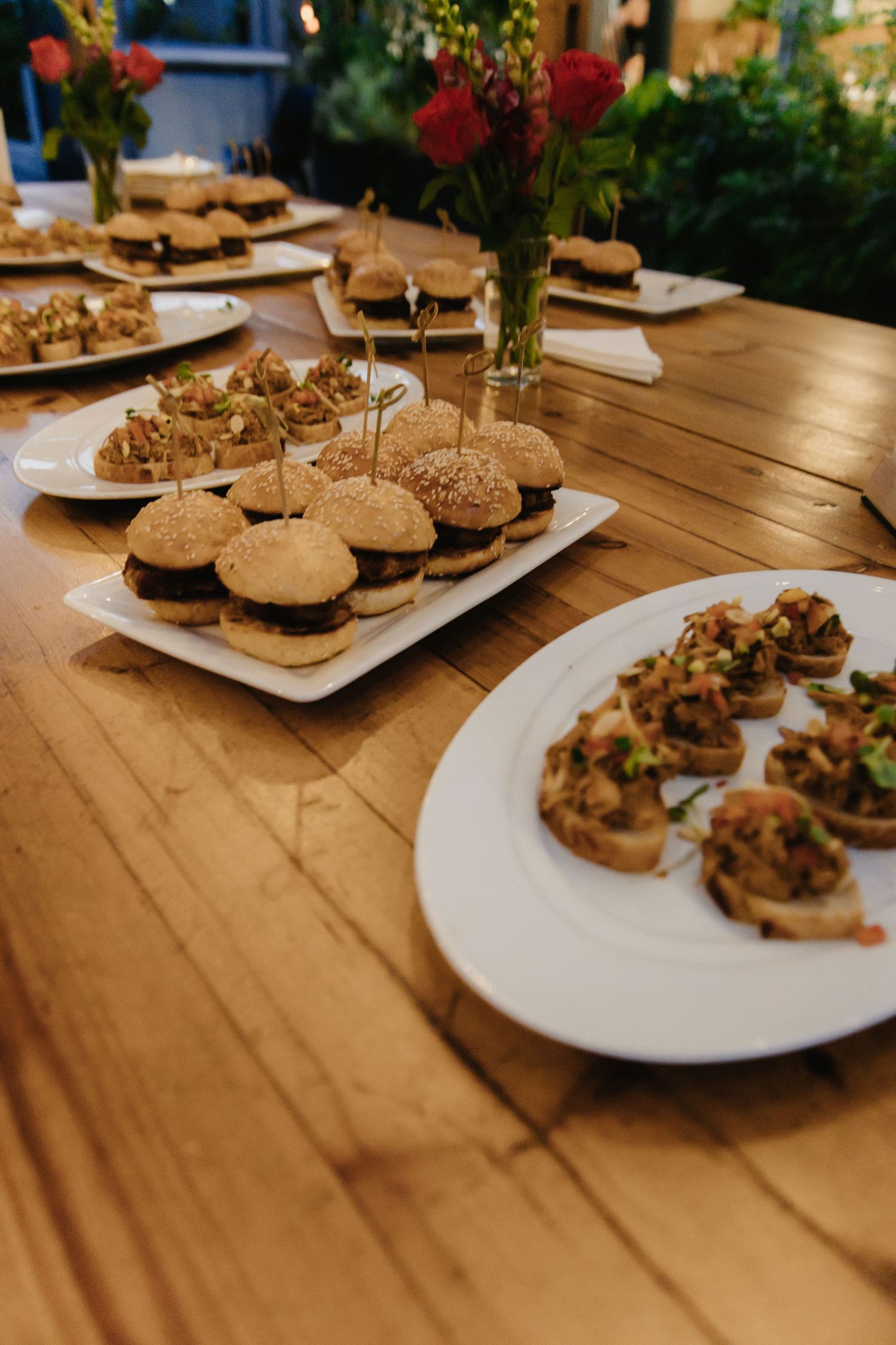 White plates of gourmet sliders and appetizers arranged on a rustic wooden table at a catered event.