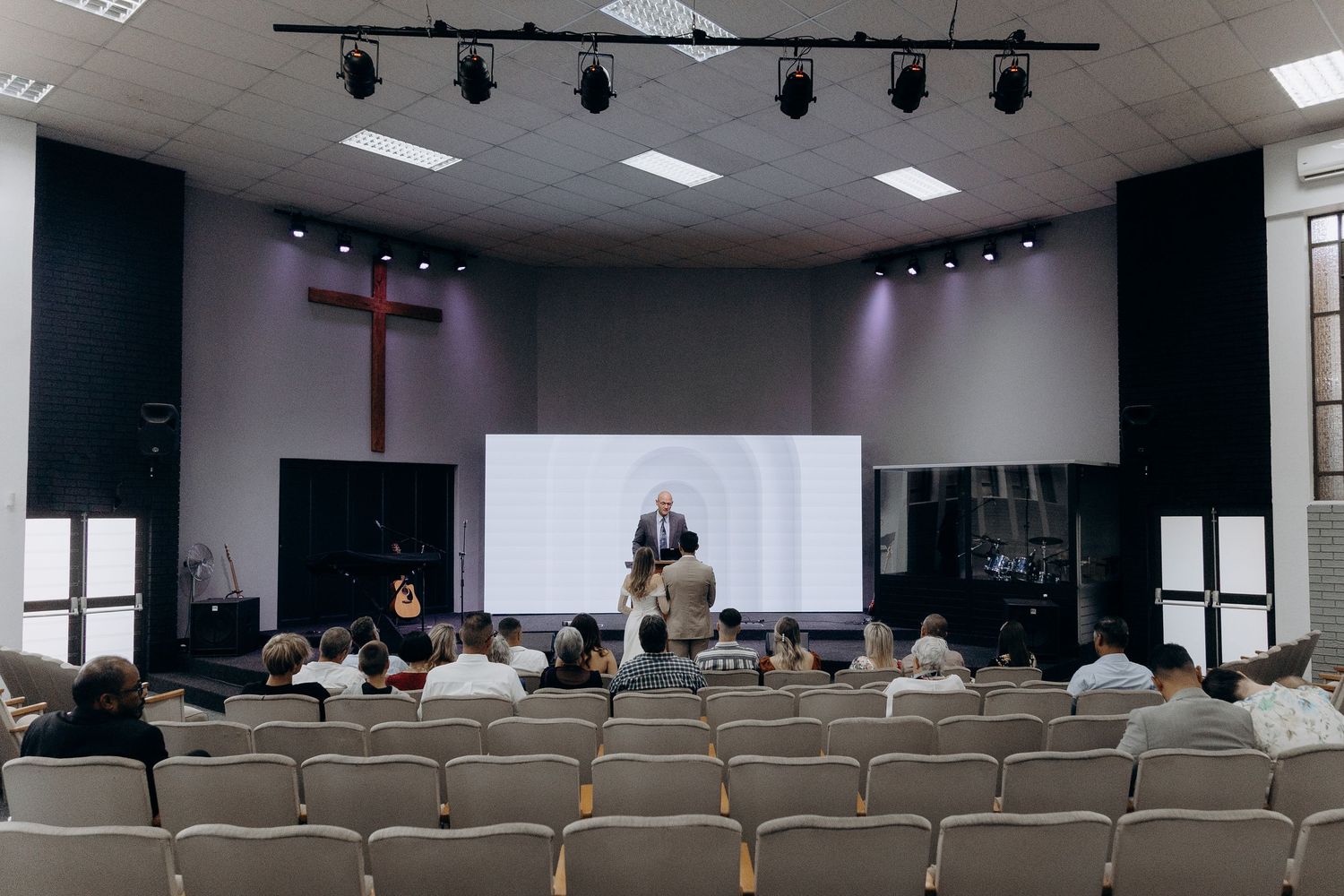 A church sanctuary with white chairs and stage lighting features a presentation screen and cross on the wall.
